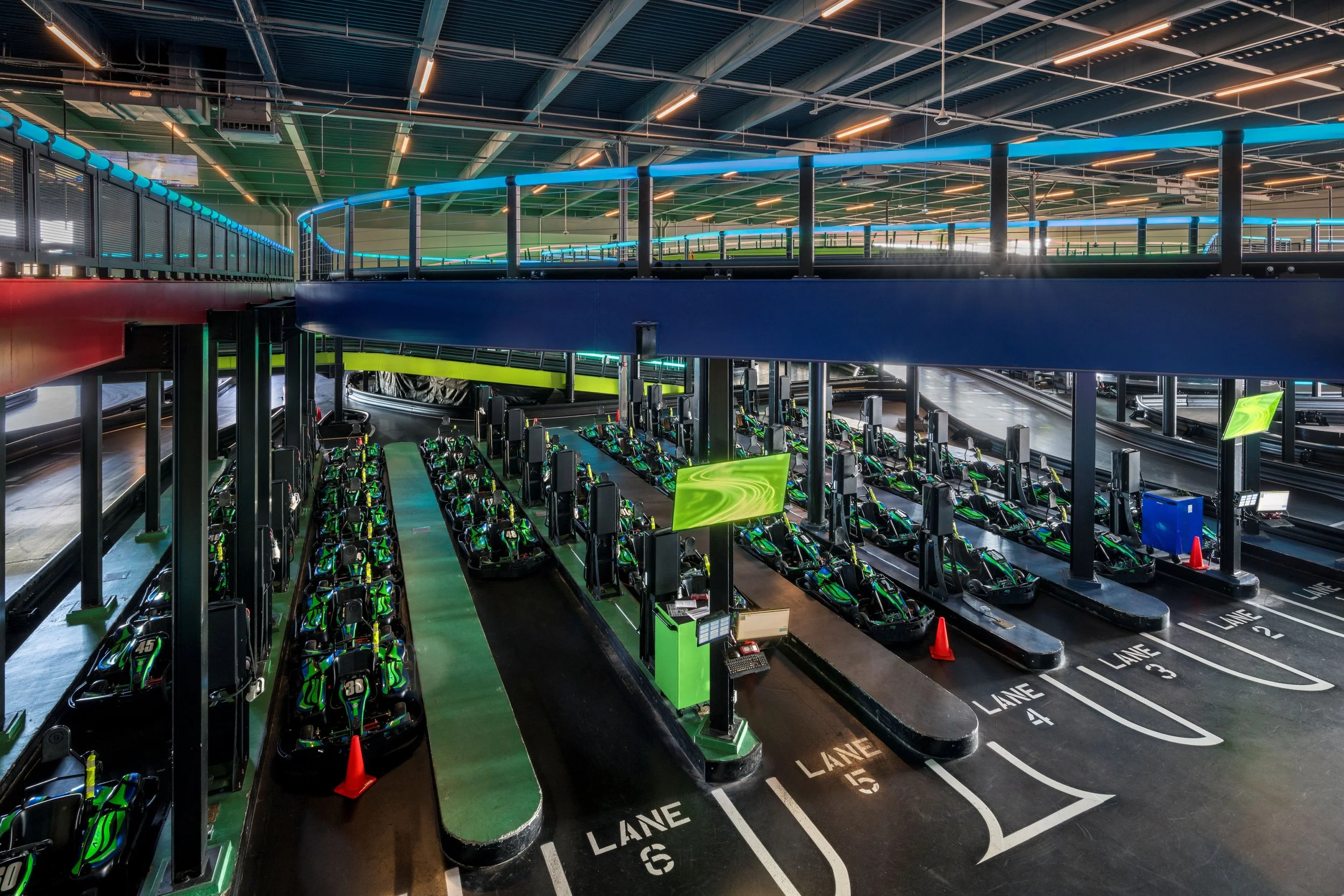 Indoor go-kart racing track with multiple go-karts parked in designated lanes, illuminated by colorful neon lights, and a second level viewing area with colorful lighting.