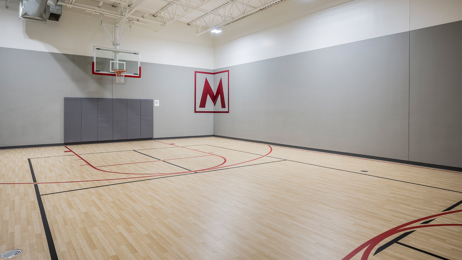 An indoor basketball court with a wooden floor, a basketball hoop, and a large capital 'M' logo on the wall.