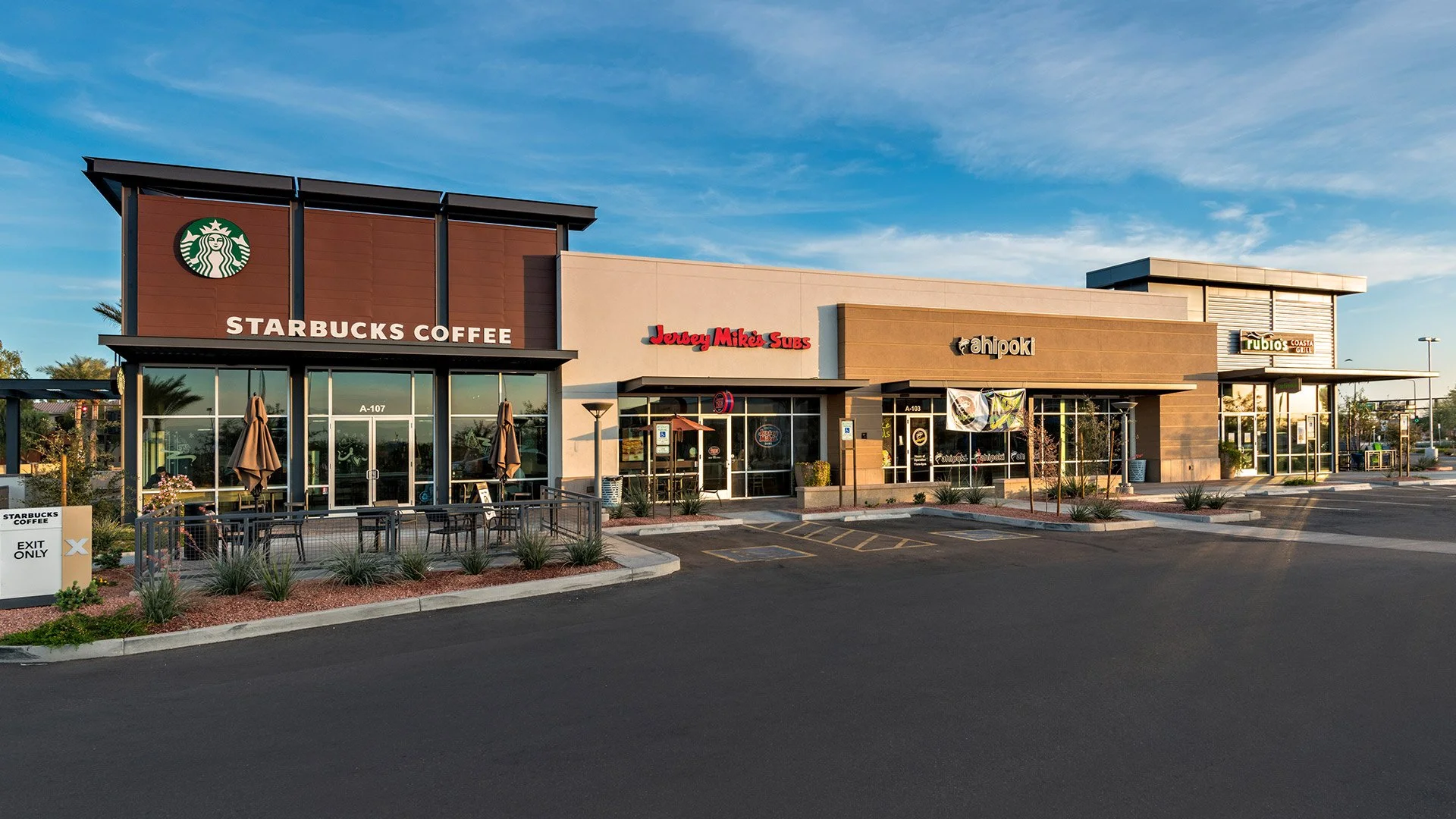 Exterior of a shopping center with a Starbucks Coffee, Jersey Mike's Subs, Ah Poki, and Rubios Coastal Grill, parking lot in front, clear sky