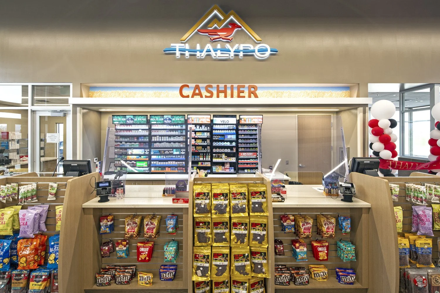 Supermarket checkout counter with candy and snack displays, cash register, and a sign reading 'CASHIER'. Behind the counter are shelves with tobacco products, a credit card reader, and a computer. Balloon decorations are visible on the right side.
