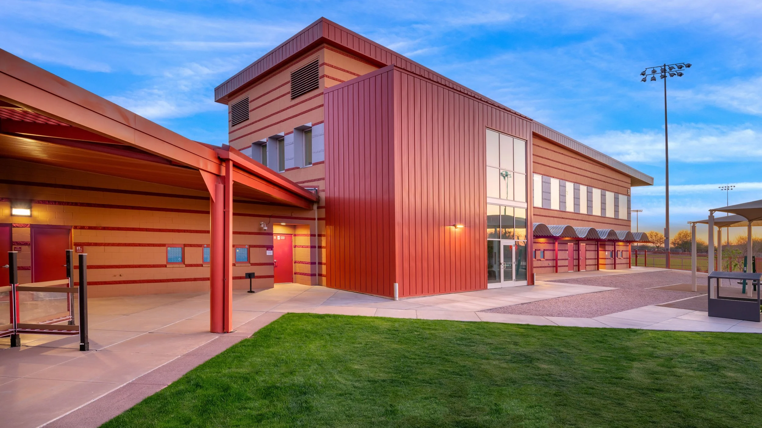 Exterior view of a modern building with a red and beige color scheme, large glass windows, and a grassy area in the foreground at sunset.