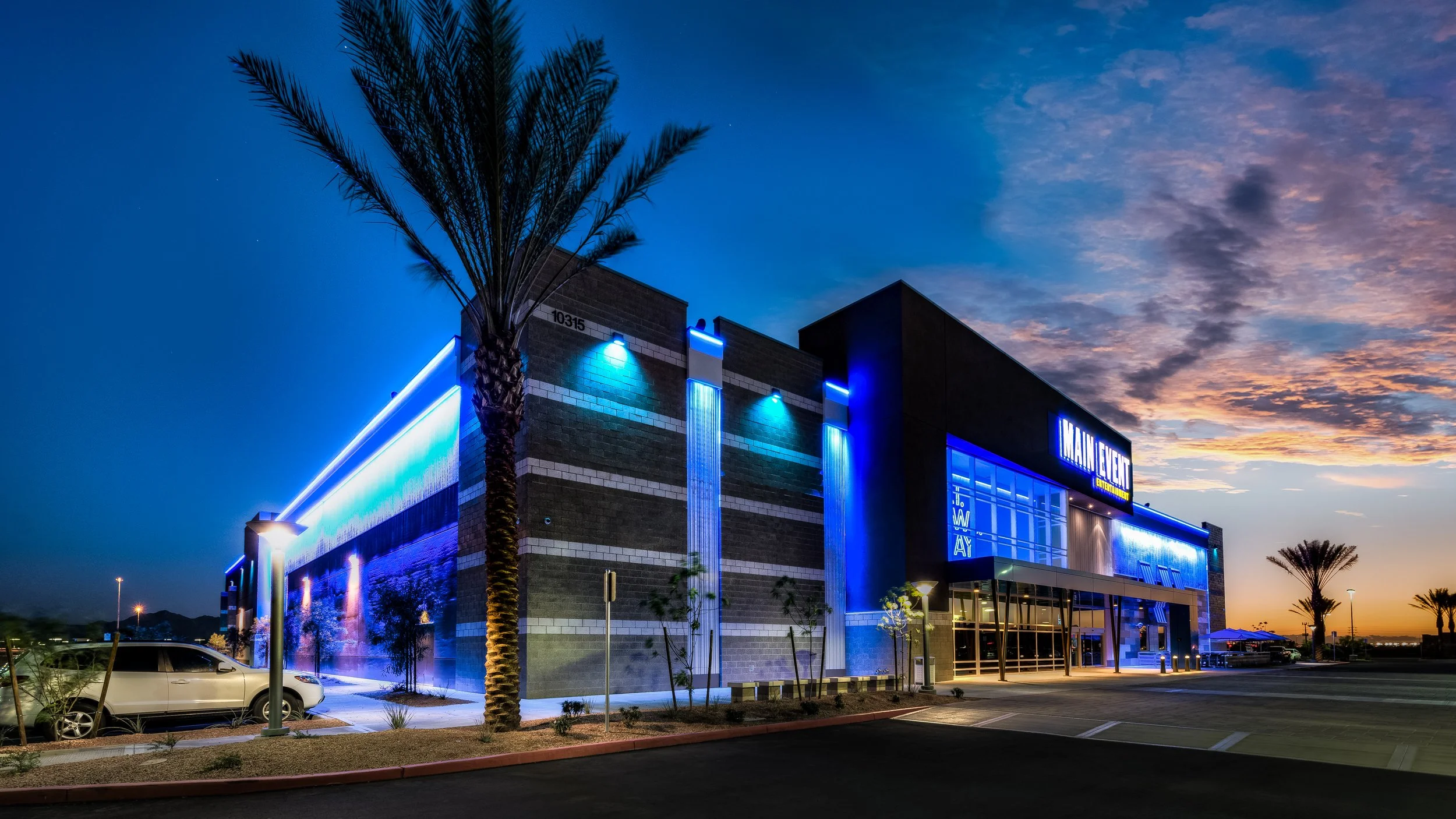 Exterior of an entertainment venue called Main Event during twilight with blue neon lighting, palm trees, parked cars, and a partly cloudy sky.