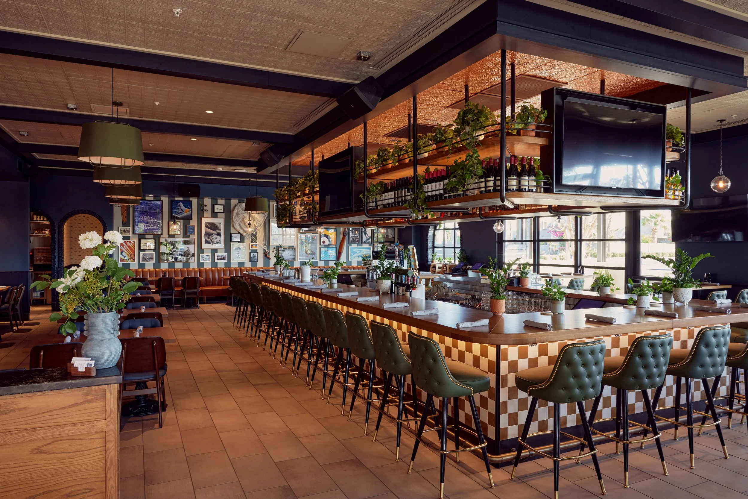 Interior of a stylish restaurant with a bar counter, green chairs, and decorative plants, featuring a gallery wall with various artworks.