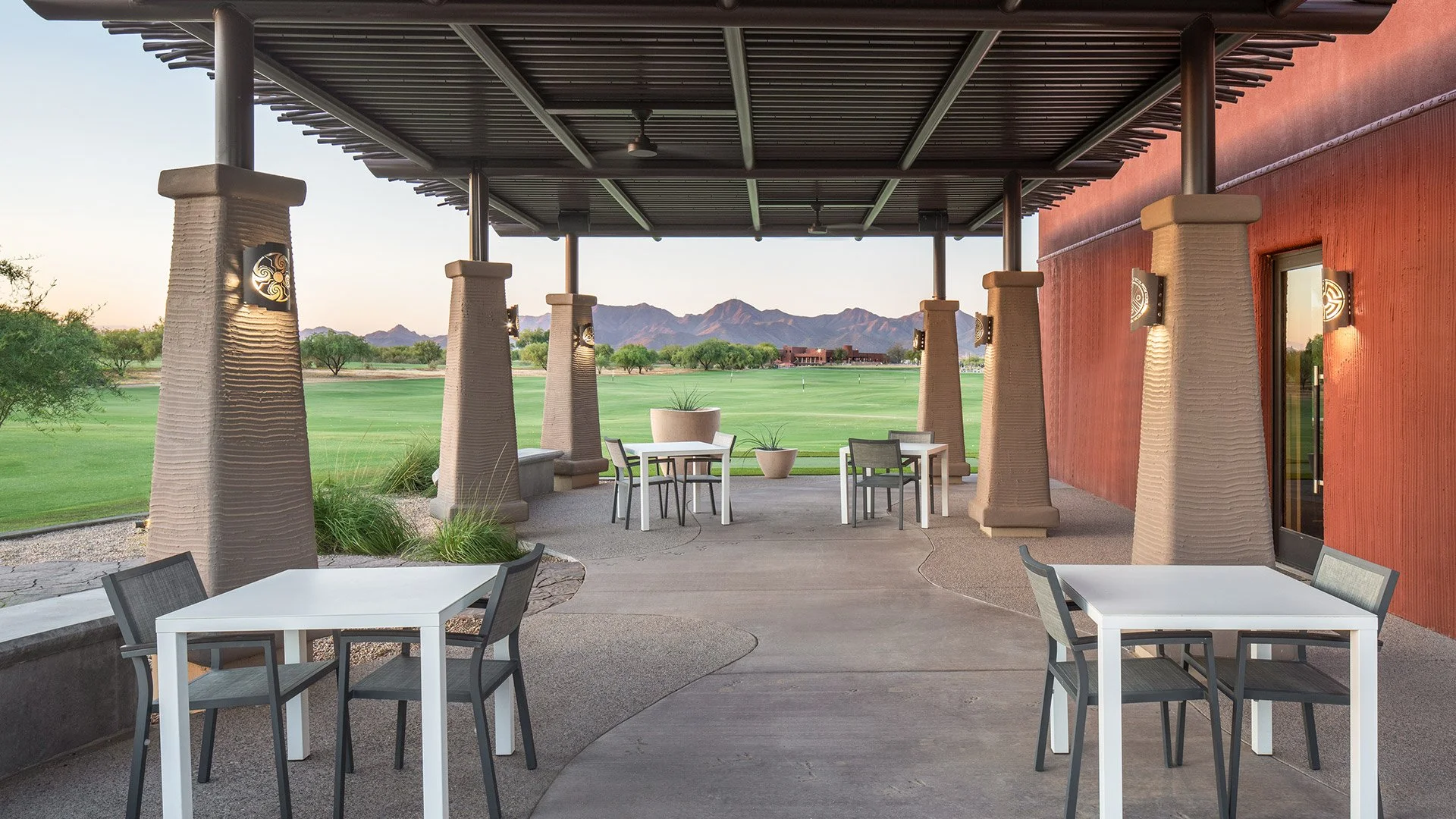 Outdoor patio with white tables and black chairs, stone pillars, green grass golf course, mountains in the background, red building exterior, potted plants, and sunset lighting.