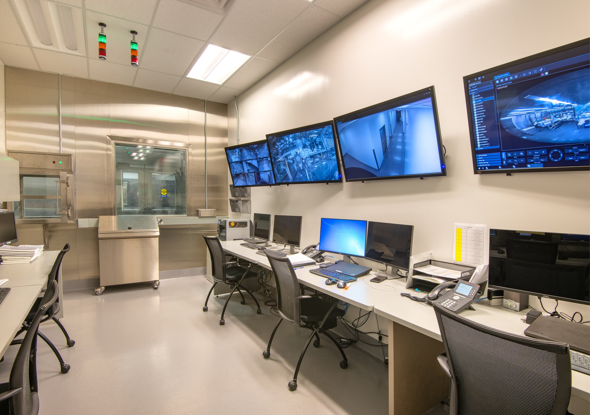 Control room with multiple computer monitors displaying security camera feeds, a window into a sterile laboratory, and various office supplies and equipment on desks.