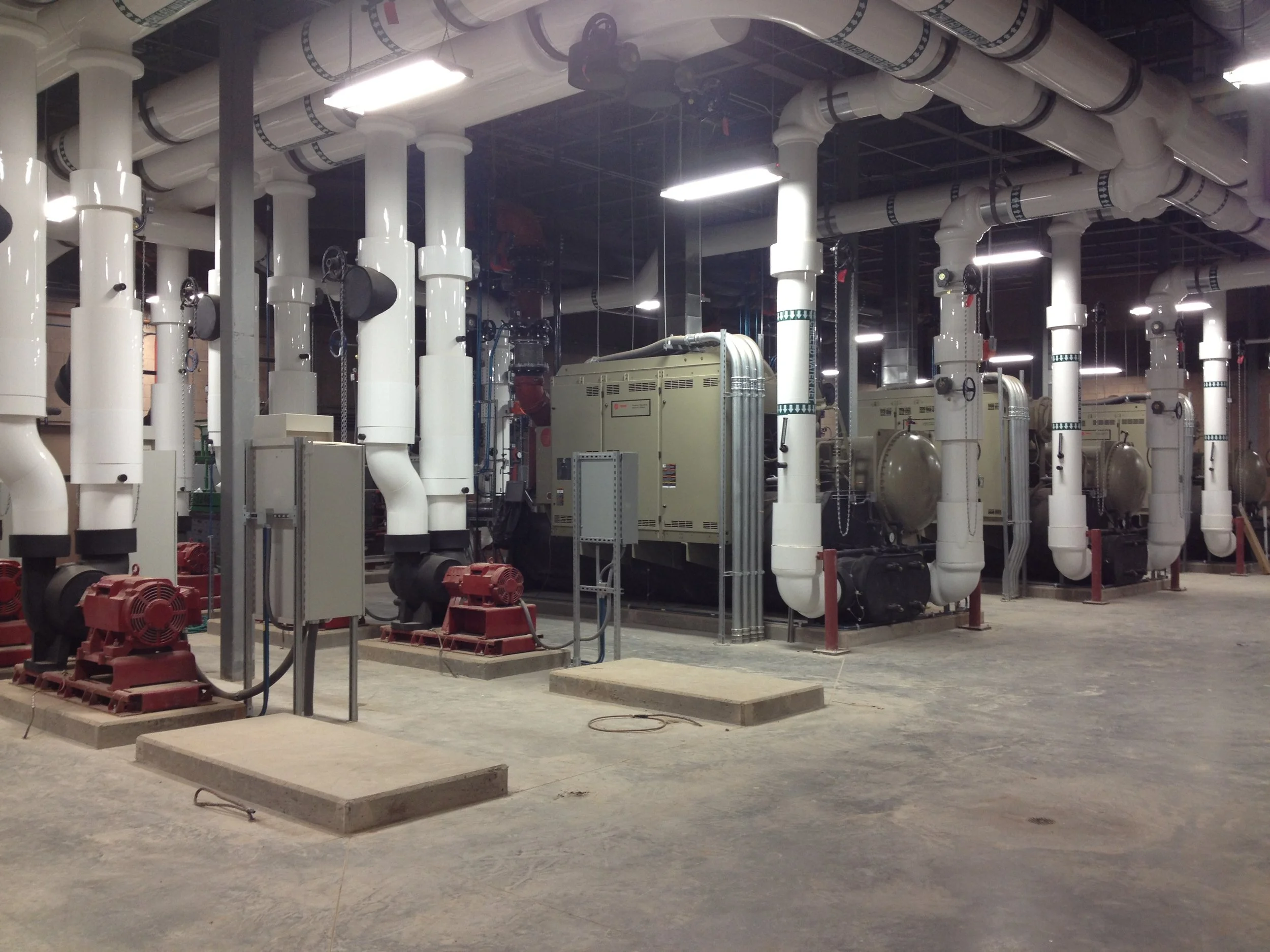 Industrial room with large white pipes, pumps, and machinery, with concrete flooring and overhead lighting.