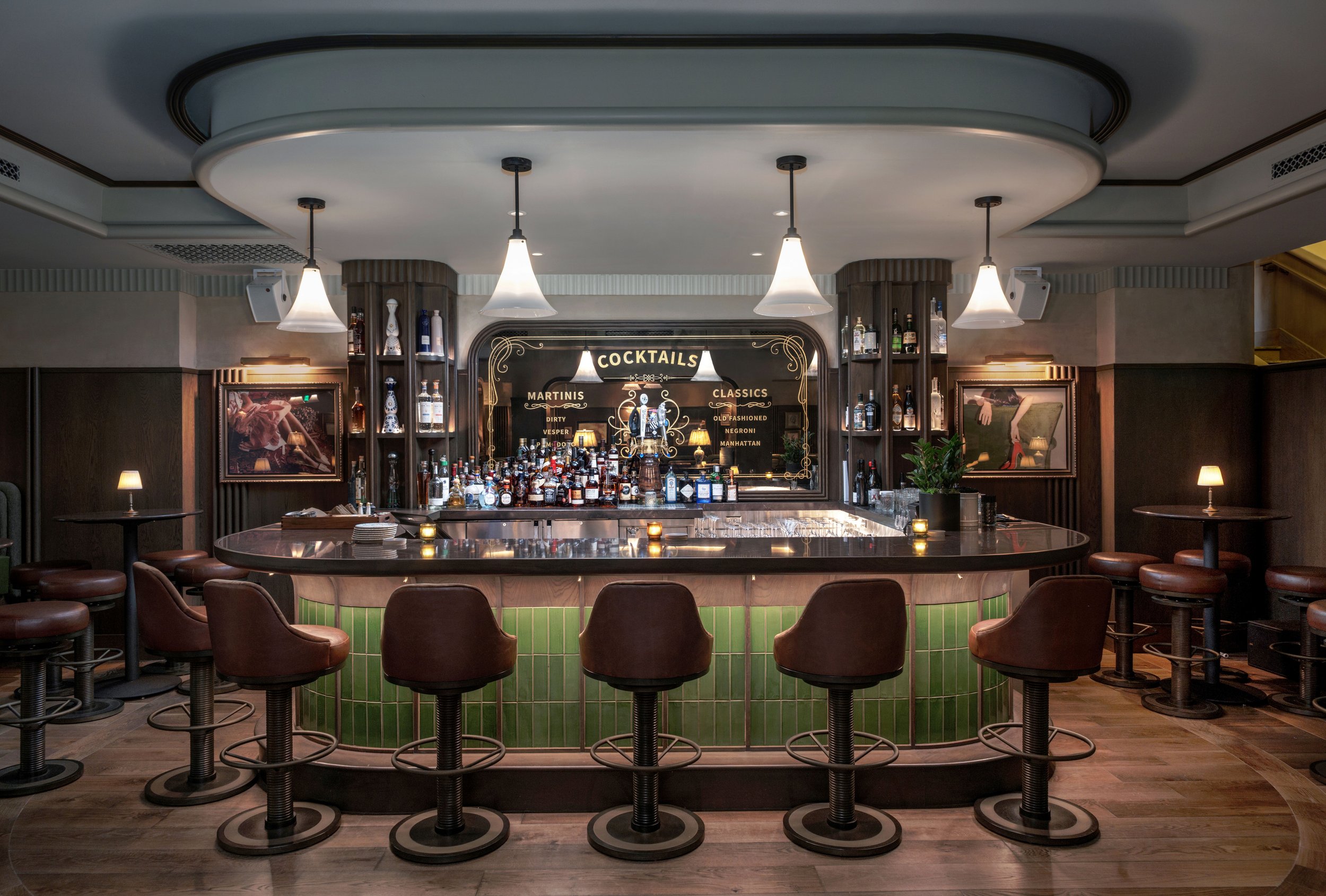 Empty bar with a black countertop, brown bar stools, liquor bottles on shelves, framed pictures on the walls, and a cocktail menu behind the bar.