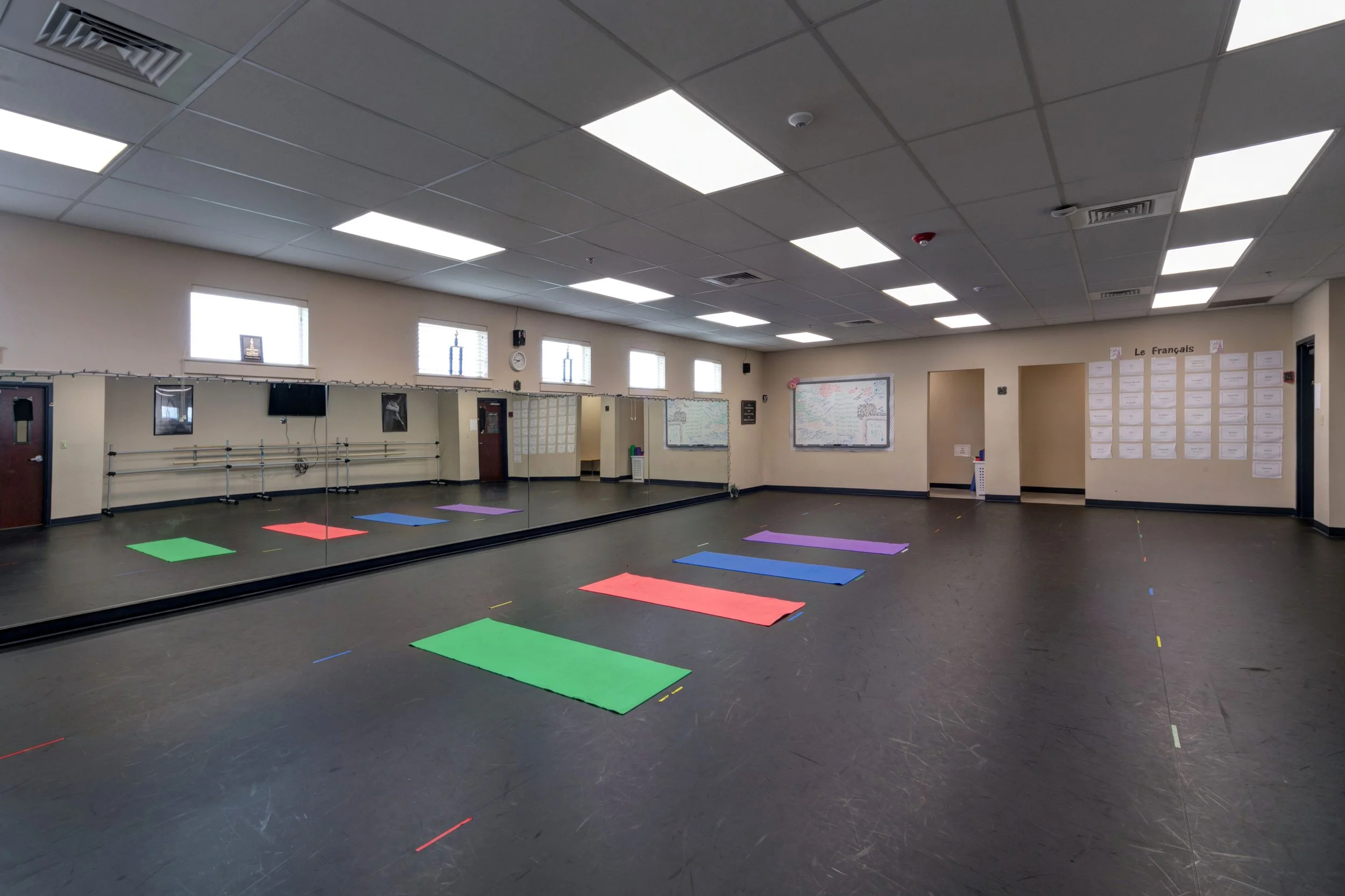 Empty dance studio or classroom with colorful mats on black floor, mirrored wall, white walls, and ceiling lights.