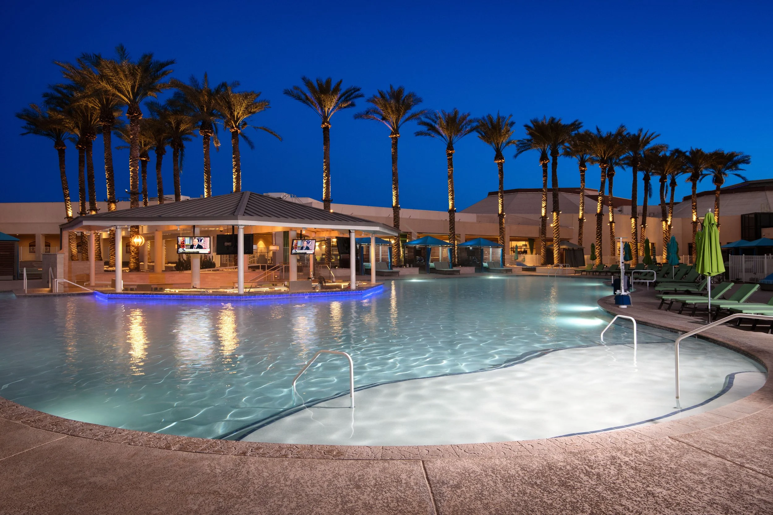 Night view of a resort pool area with palm trees, lounge chairs, umbrellas, and a pool bar with TVs, illuminated with soft lighting.