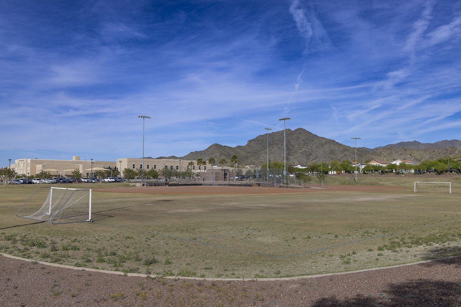 An empty sports field with two soccer goals, a parking lot, a school building, palm trees, and mountains in the background under a blue sky with wispy clouds.