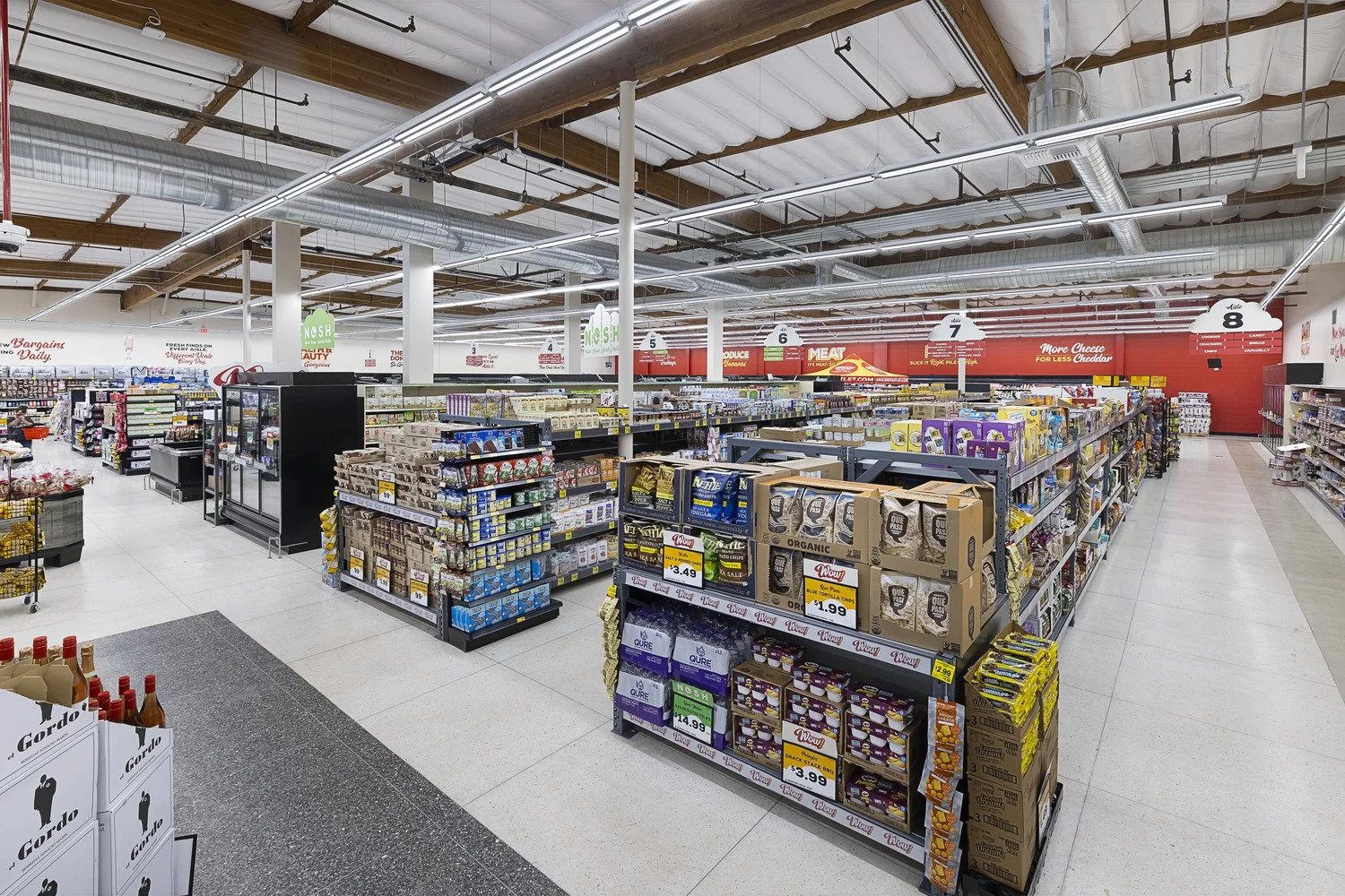 Interior of a grocery store aisle with shelves stocked with various food products, including snack packs, drinks, and canned goods. Bright overhead lighting and a clean, organized space.