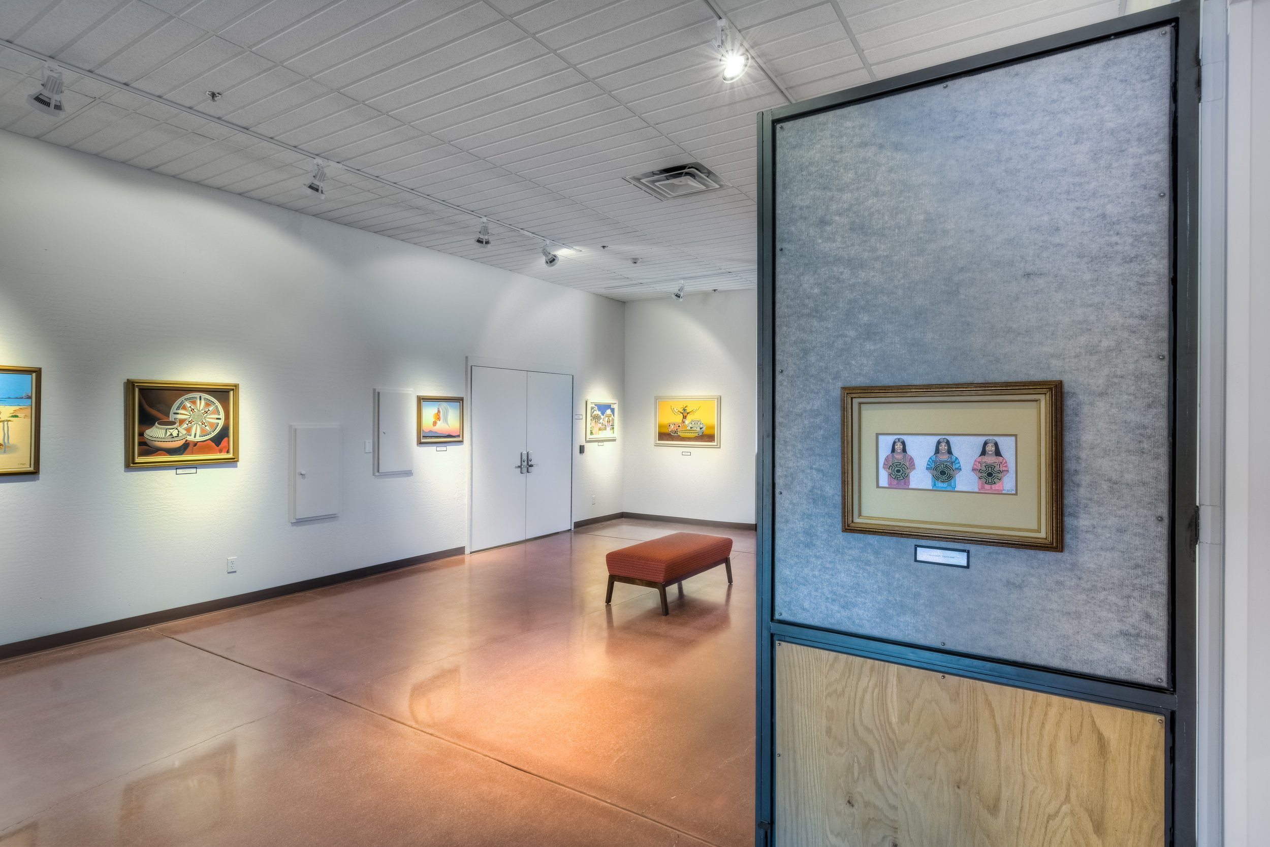 Interior view of an art gallery with paintings on white walls, a red bench in the center, and a display board with a framed artwork of three women in traditional clothing.