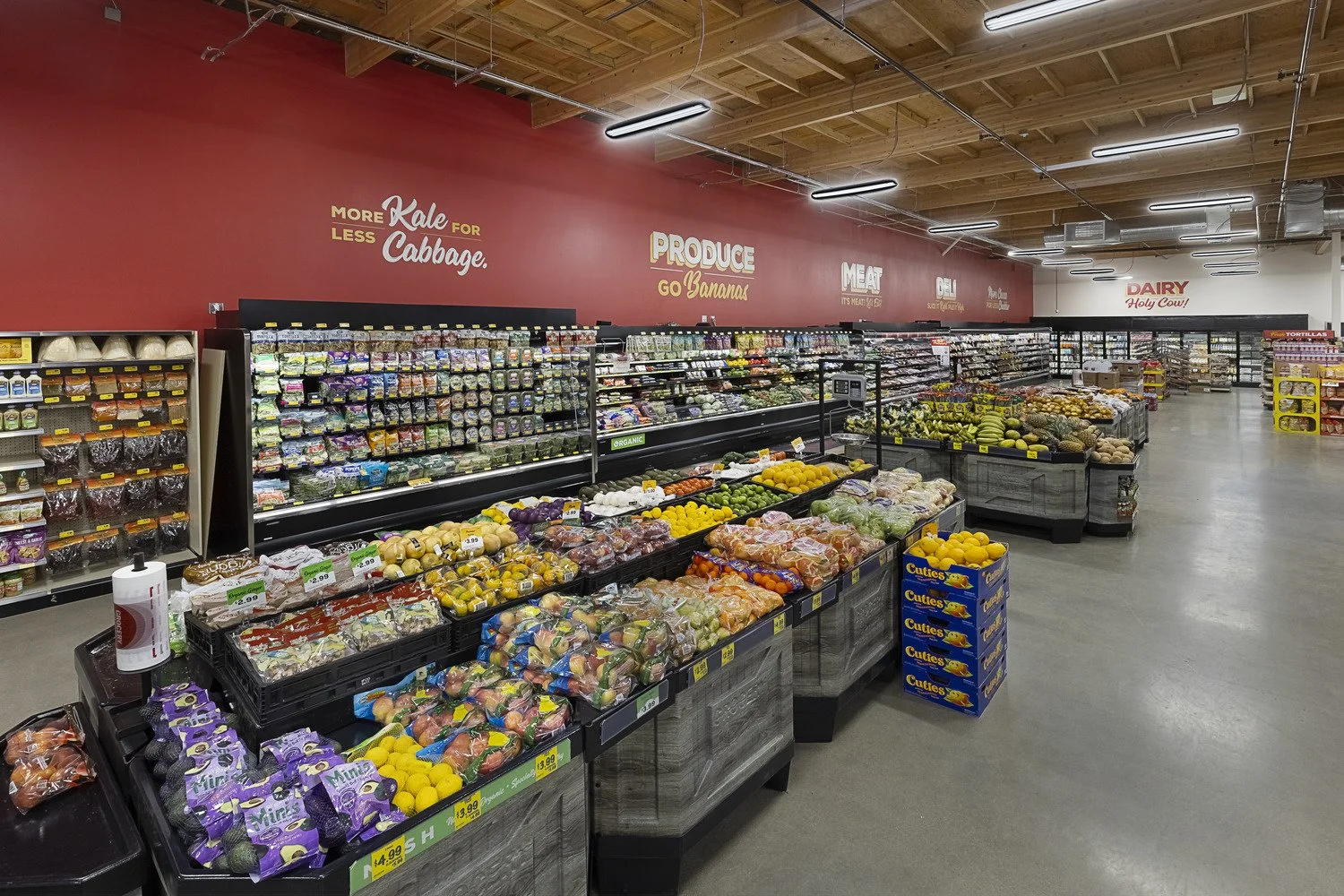 Interior of a grocery store produce section with various fruits and vegetables on display, including bananas, apples, lemons, and leafy greens, with a red wall and signage.