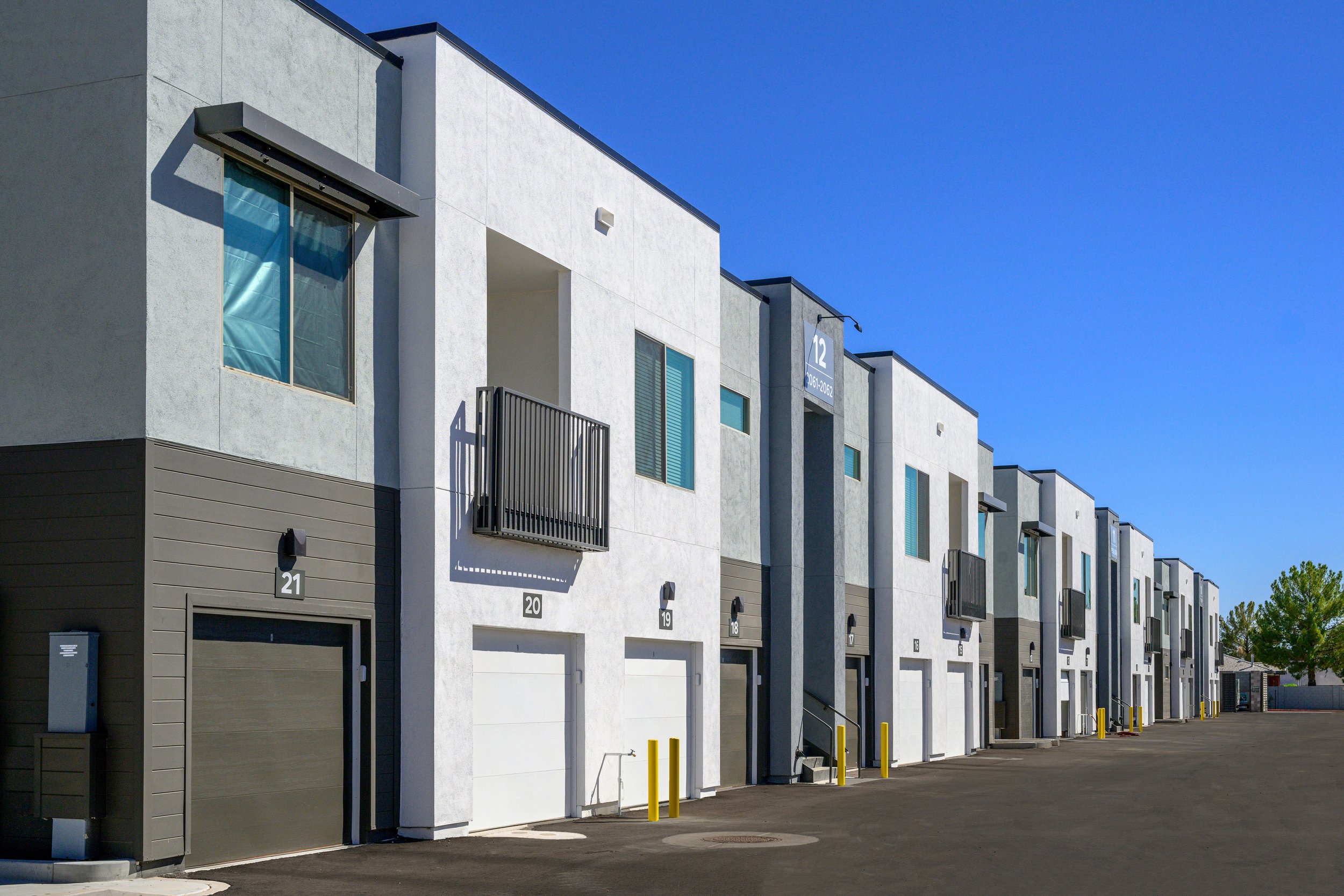 Modern white and gray multi-story apartment building with garages and small balconies, bright blue sky, and a paved parking area in front.