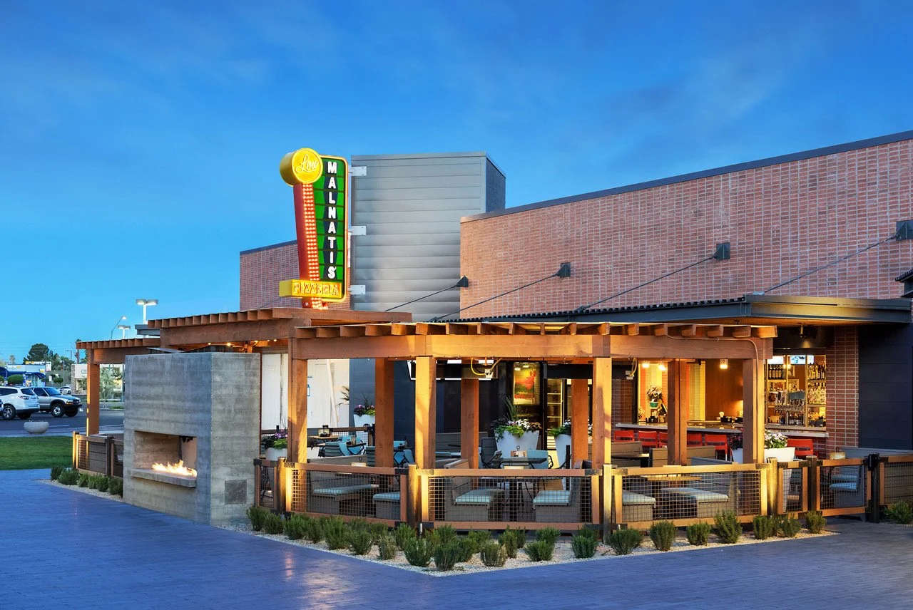 Exterior view of Low Malinas restaurant with a brick building, a wooden patio area with seating, and a neon sign.