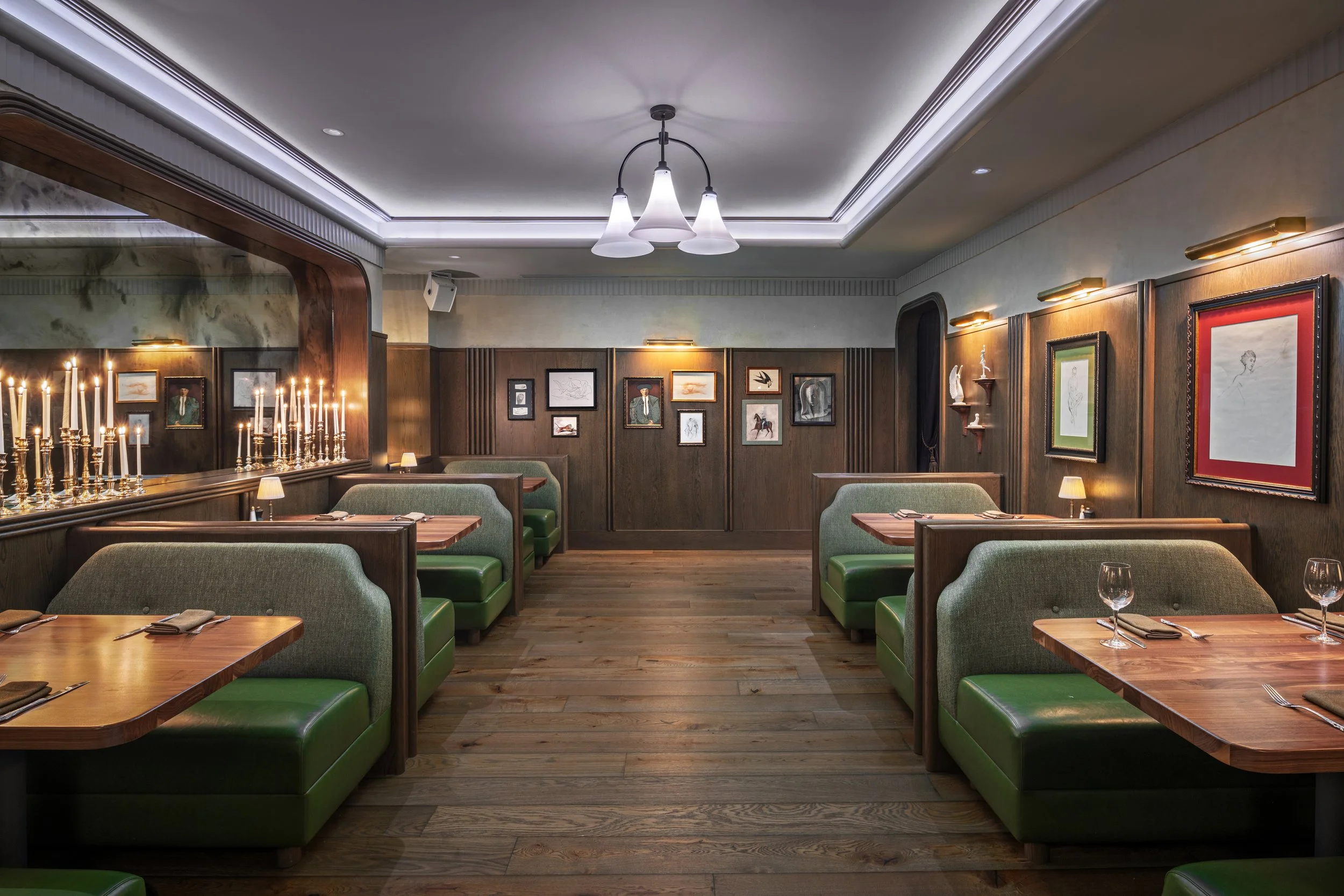 Interior of a cozy restaurant with dark wood paneling, green upholstered booths, wooden tables set for dining, and framed artwork on the walls. Candle-like lights and small table lamps create warm lighting.