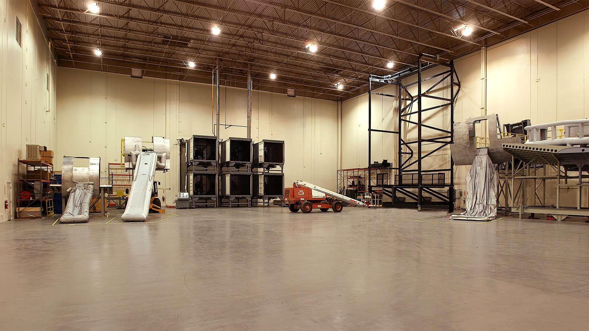 Interior of a large industrial or amusement park space with empty areas, various scaffolding, equipment, and structures, including slides and a lift platform.