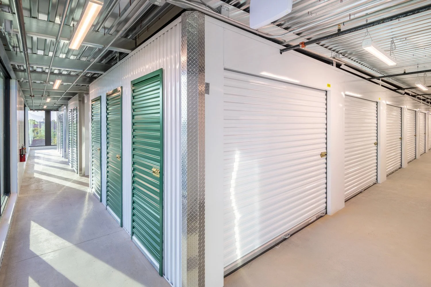 Row of storage unit doors in a corridor, some are green metal roll-up doors, others are white roll-up doors, in a modern self-storage facility.