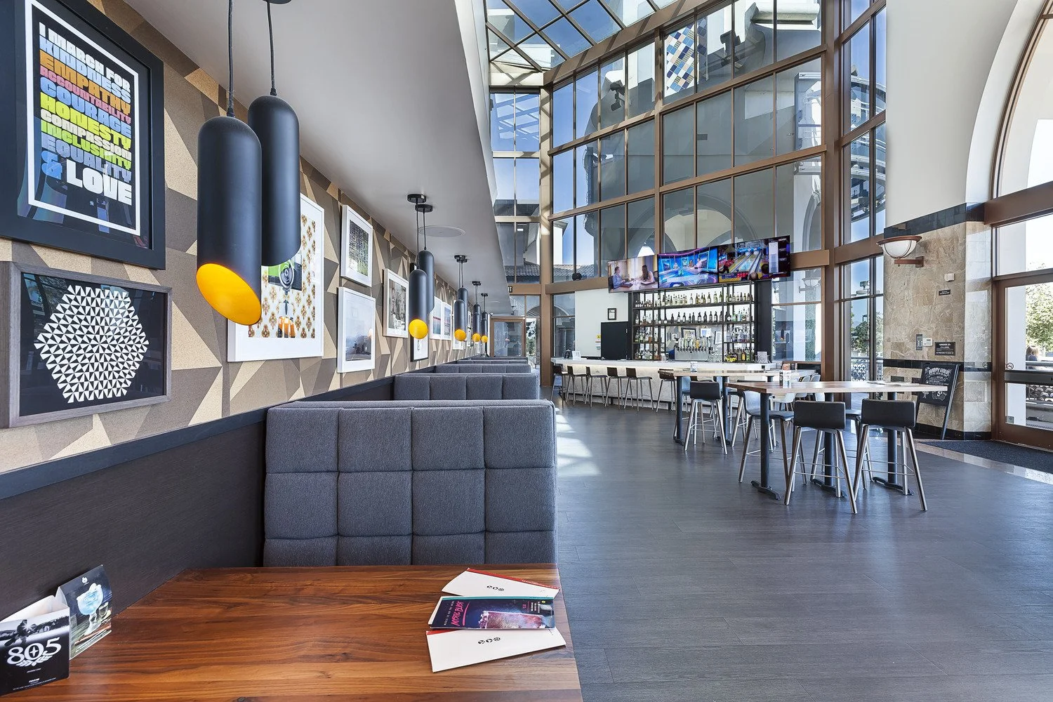 Modern, spacious bar and lounge area with large windows, contemporary wall art, black pendant lighting, tables, chairs, and a well-stocked bar with multiple TV screens on the wall.