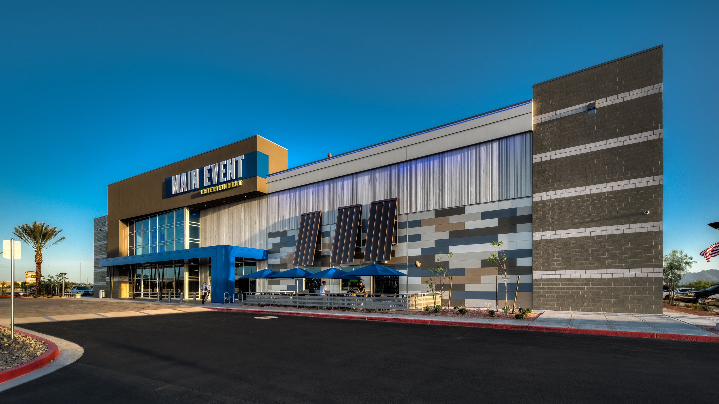 Exterior of Main Event entertainment center with a modern facade, blue accents, umbrellas outside, and a clear blue sky.