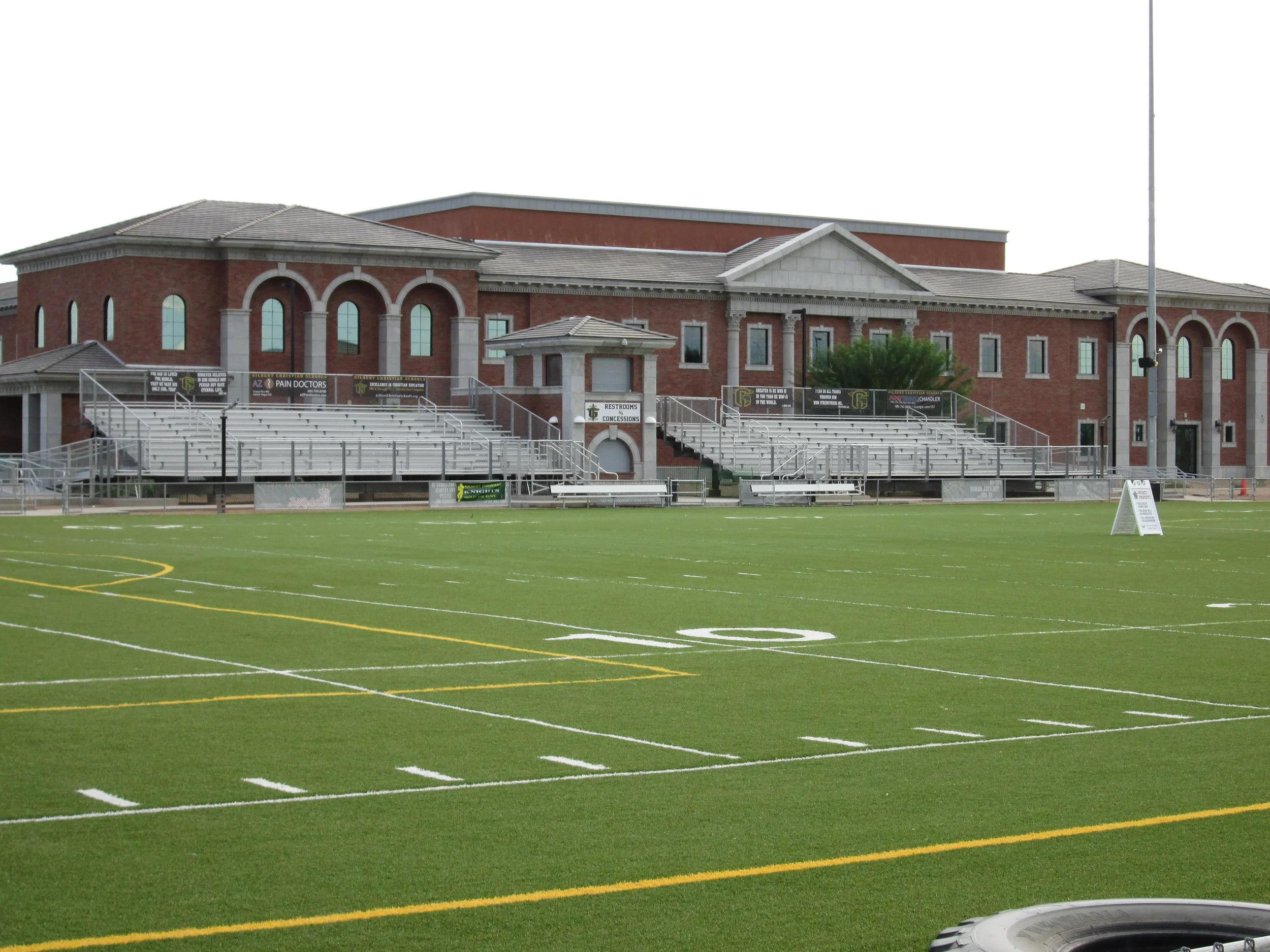Empty sports stadium with green artificial turf field, metal bleachers, brick building with arched windows in the background, and a sign on the field.