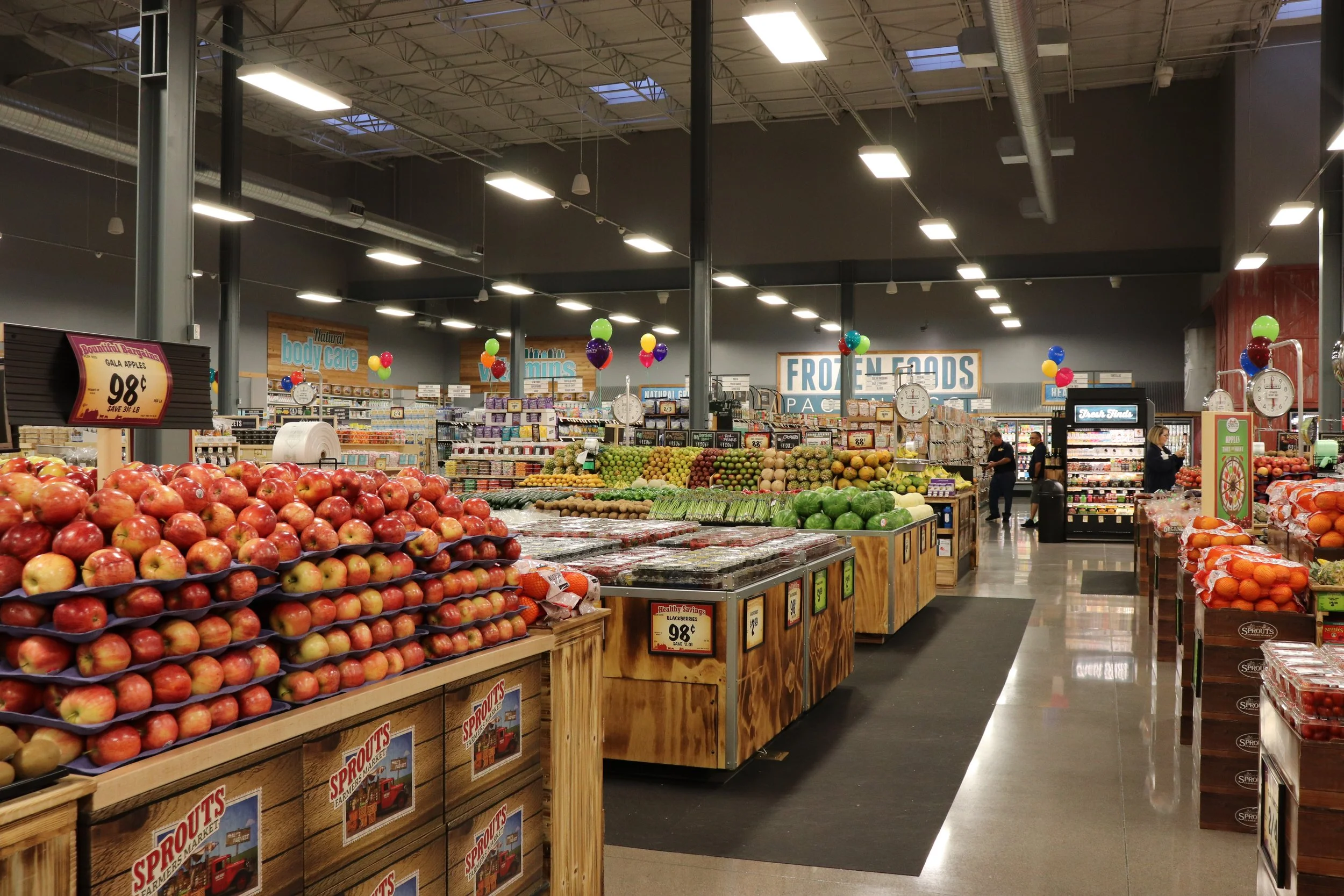 Interior of a grocery store with fresh produce stands, colorful balloons, and signs indicating sections like body care and frozen foods.