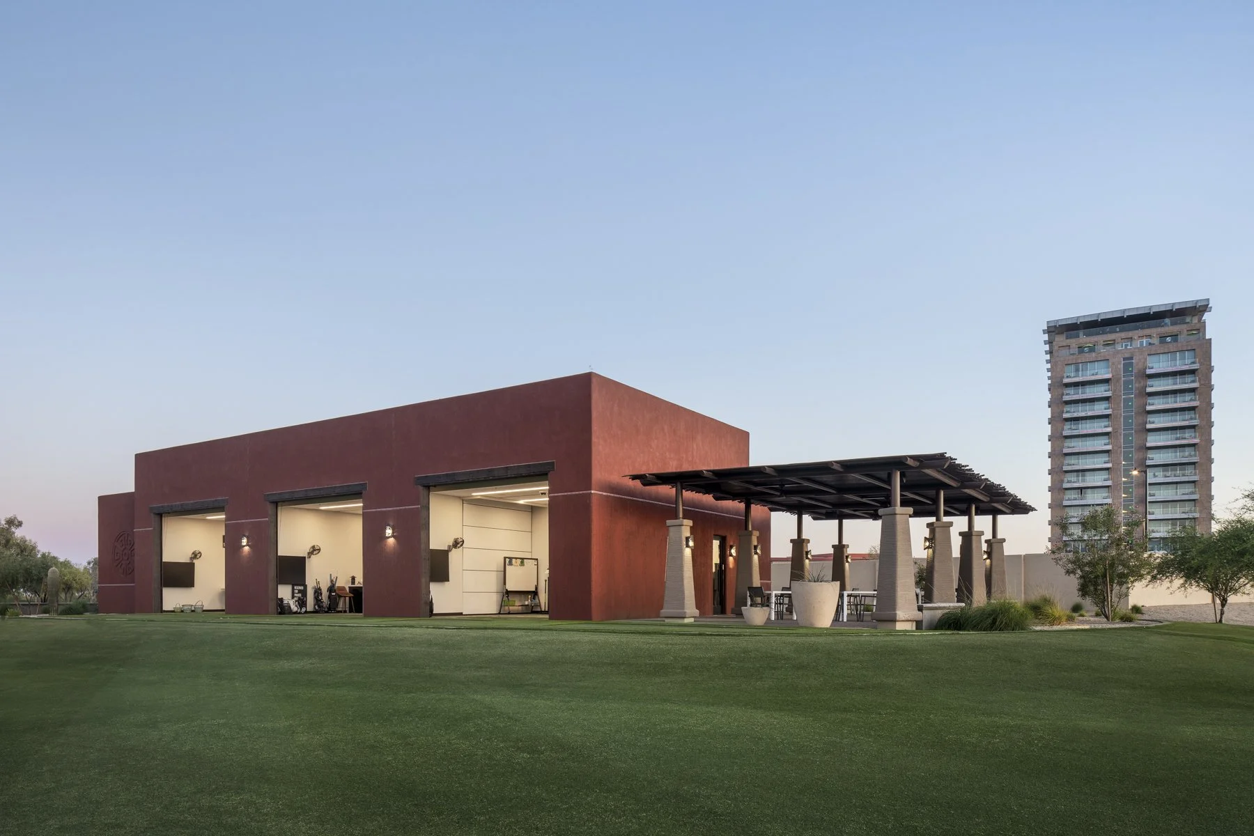 A modern red building with three large open garage-style doors, outdoor seating area with a pergola, and a tall residential building in the background during early evening.
