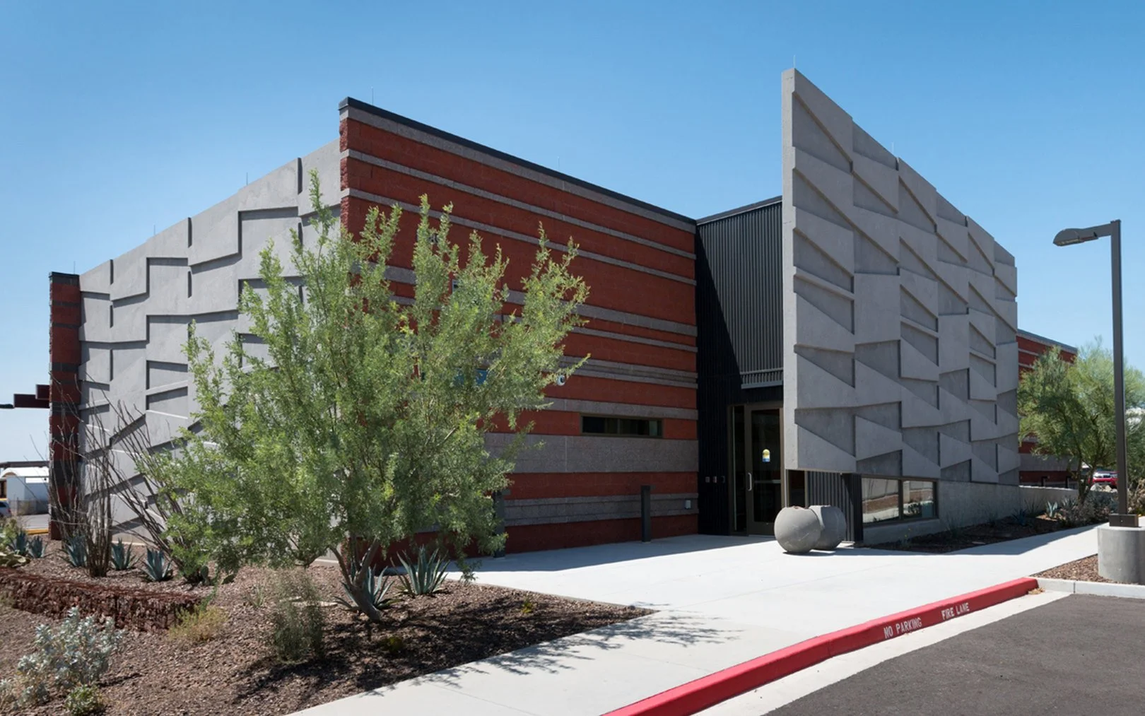 Modern building with geometric concrete and brick facade, surrounded by desert plants, with a sidewalk and a street marked 'No Parking Fire Lane' in front.