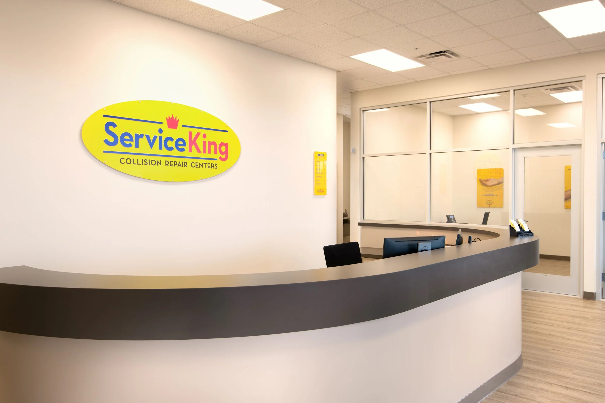 Interior of a Service King collision repair center with a reception desk, yellow Service King sign on the wall, and a windowed office area in the background.