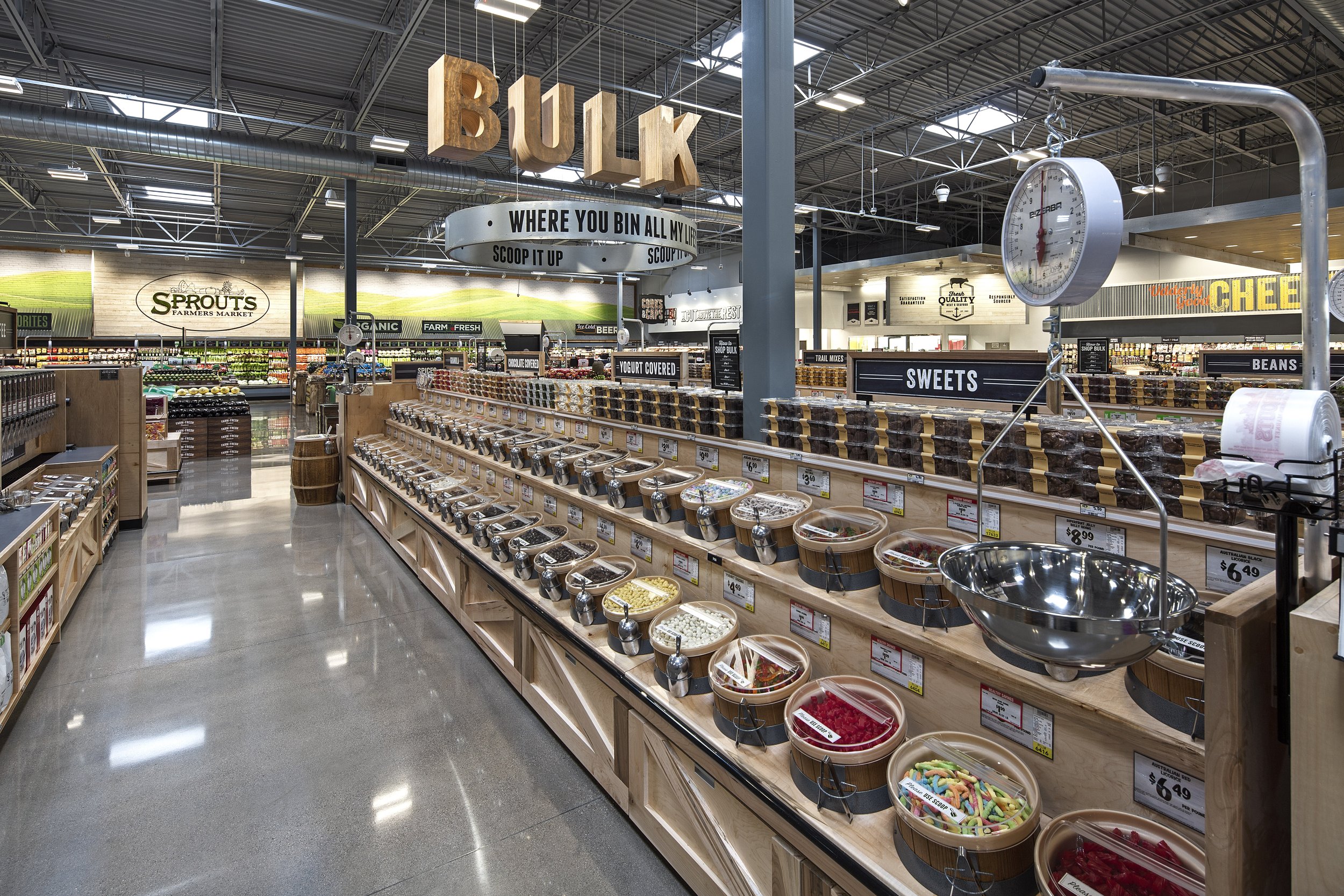 Inside a grocery store with bulk candy and other products on display. Large wooden sign reading 'BULK' hangs from the ceiling, and a scale is also visible.