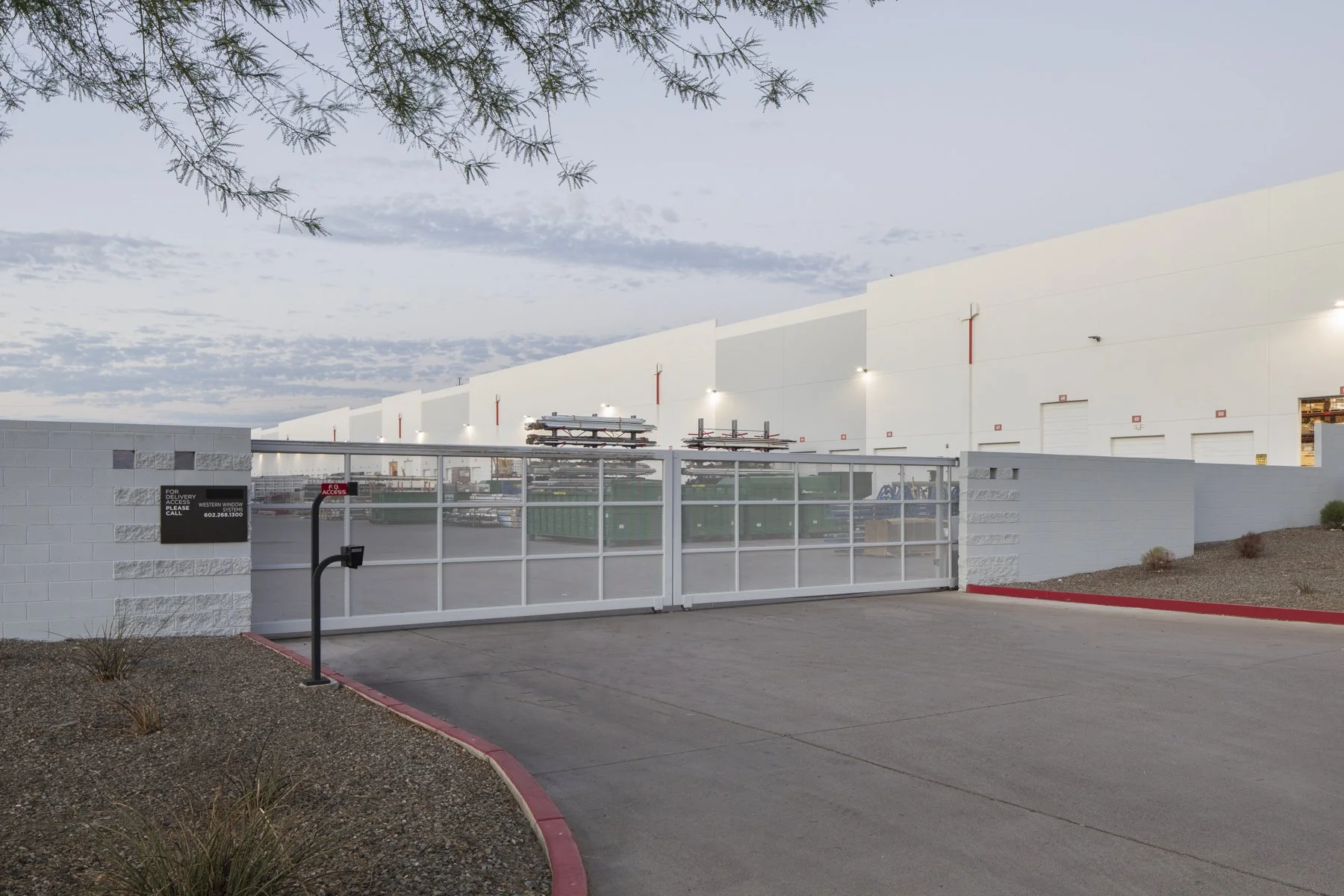 Gated entrance to a large industrial warehouse with white walls and multiple loading docks, surrounded by desert landscaping and a cloudy sky.