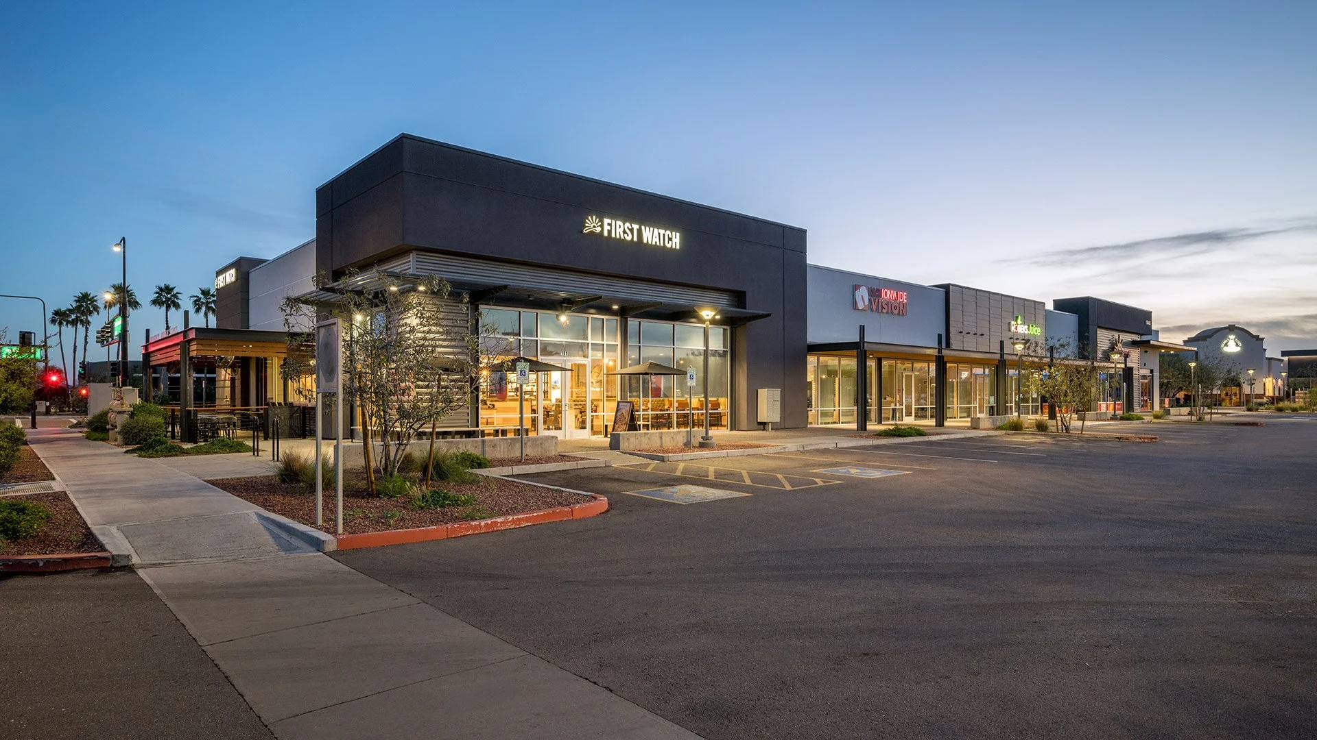 Empty shopping mall parking lot at dusk with stores including First Watch and It’s Fire visible.