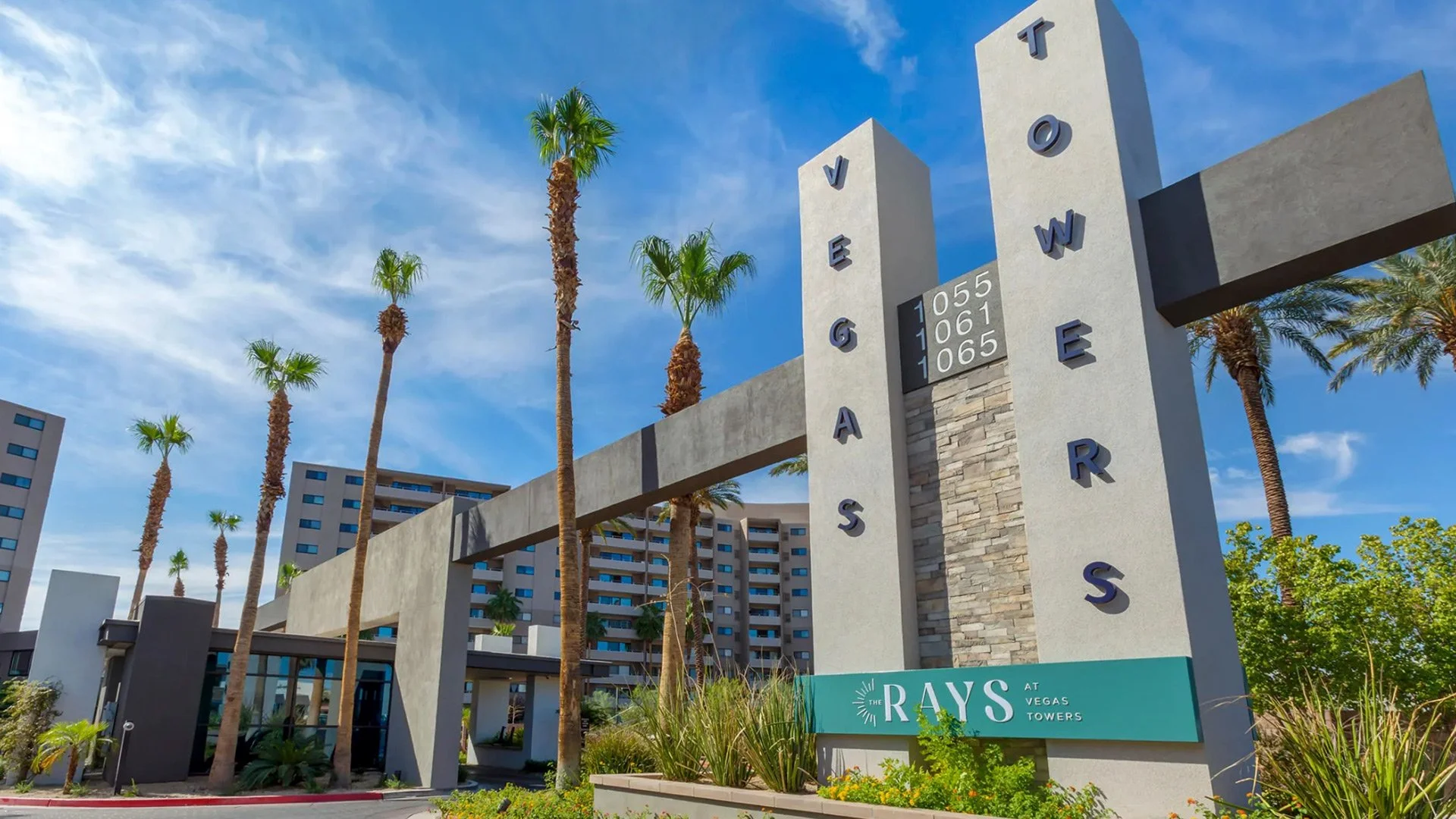 Entrance sign for Vegas Gardens at Vegas Towers surrounded by palm trees and apartment buildings under a blue sky.