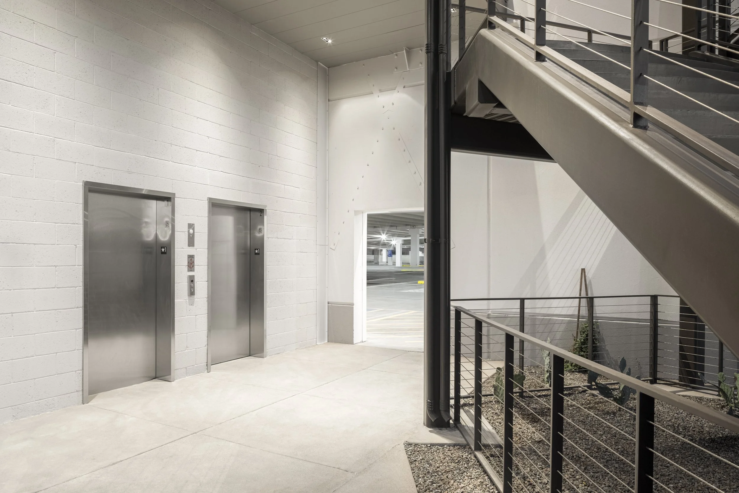 Two elevator doors in a modern indoor setting with a white brick wall and a staircase to the right, overlooking a parking garage.