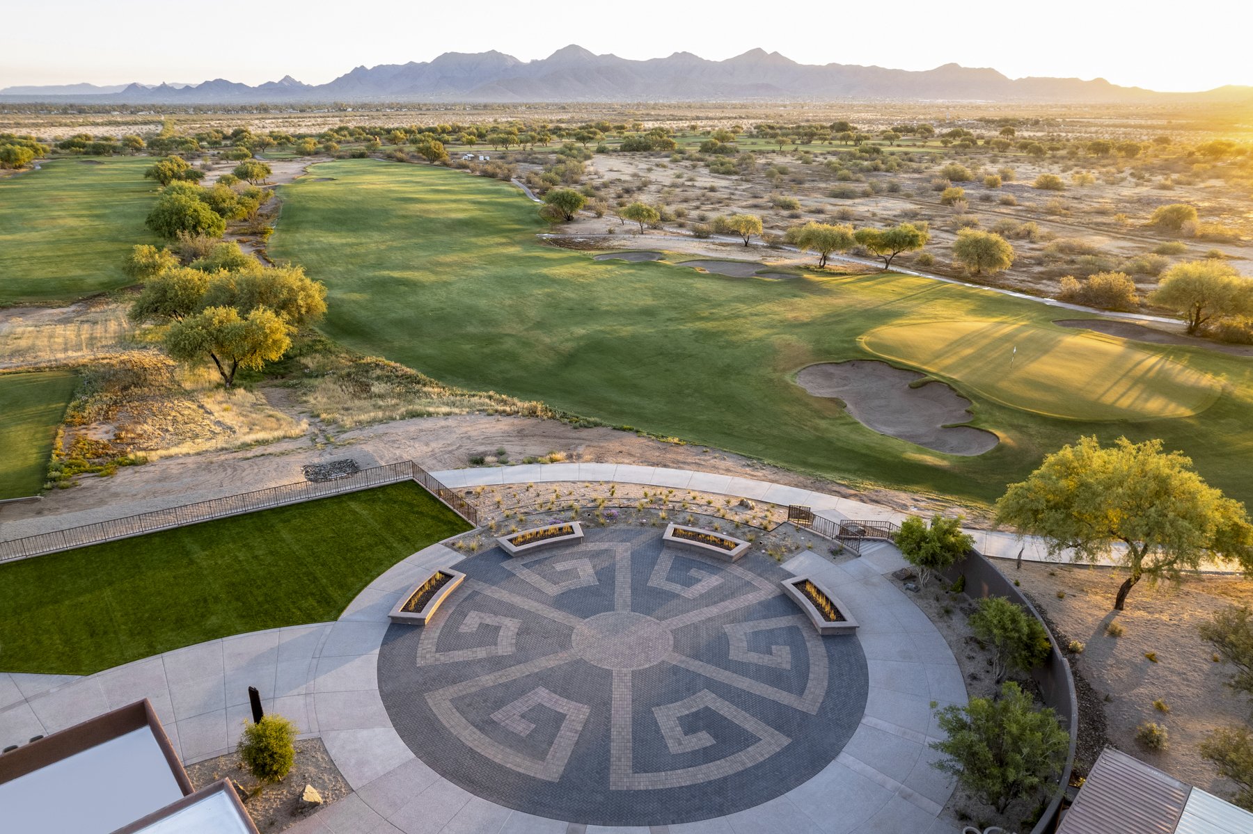 View of a golf course during sunset with mountains in the background, featuring green fairways, sand bunkers, and scattered trees.