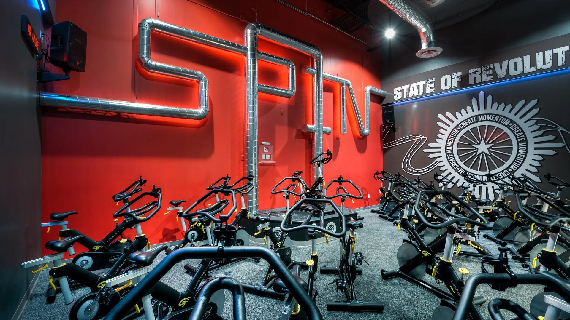 Indoor cycling studio with multiple stationary bikes arranged in rows facing a red wall with decorative metallic pipes forming the word 'SPIN' and a mural that reads 'State of Revolution' and motivational phrases.