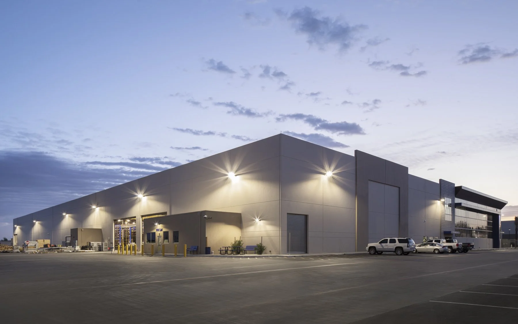 Large warehouse building with exterior lighting and parked cars, under a partly cloudy sky at dusk.