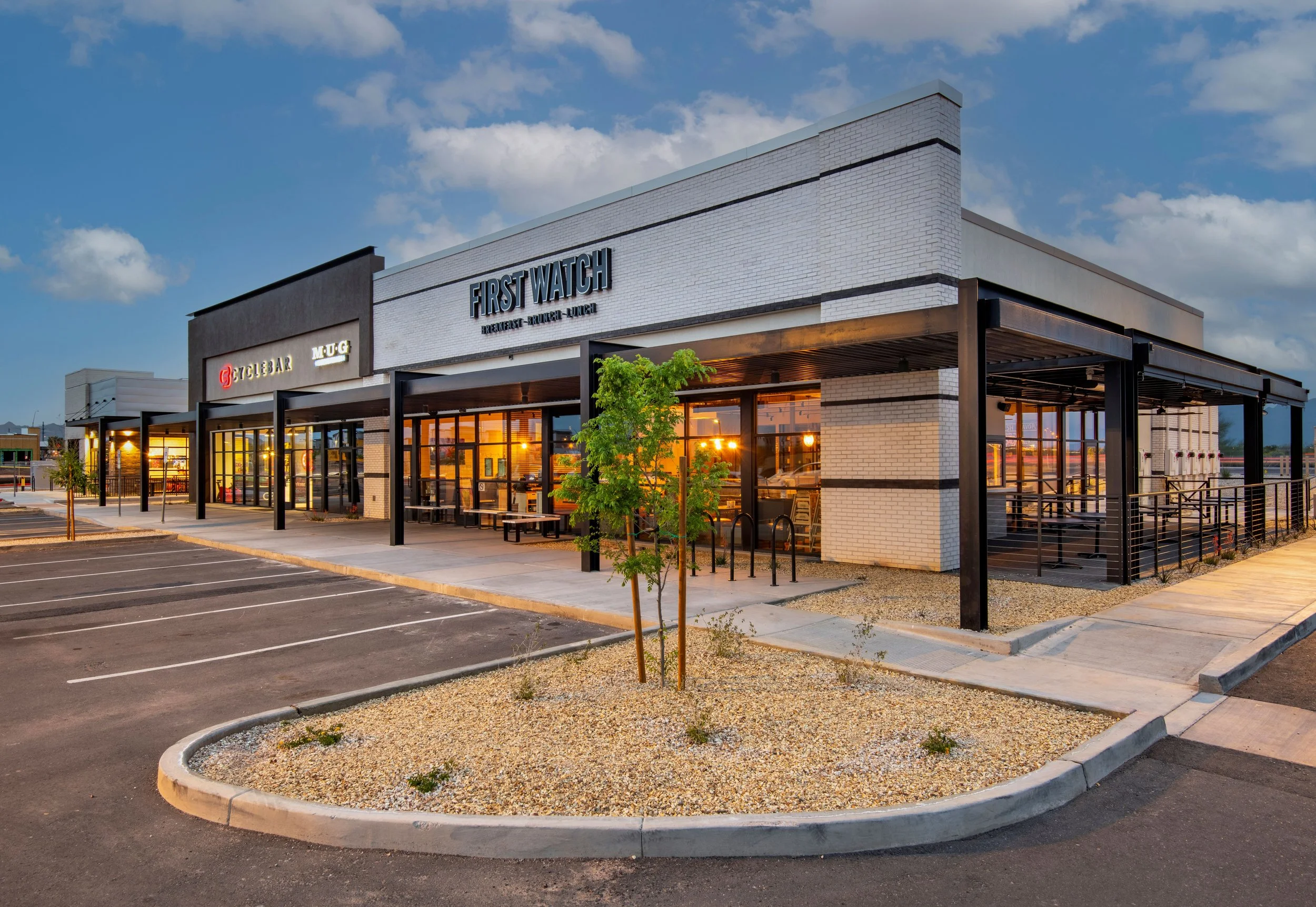 Exterior view of a modern restaurant and convenience store with seating area, parking lot, and newly planted trees at sunset