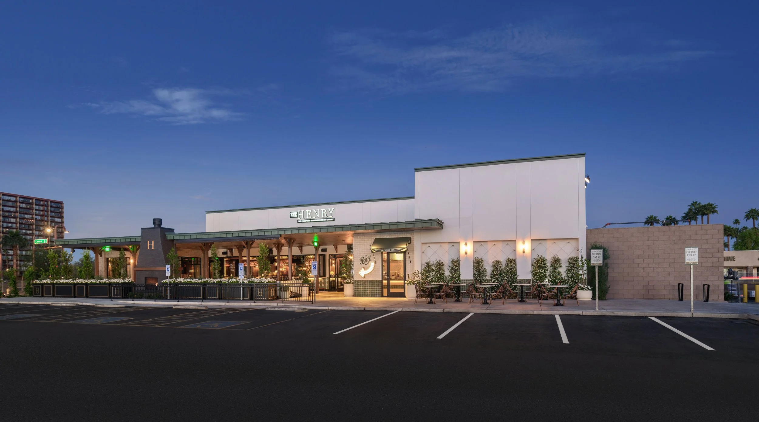 Exterior of a restaurant called HENRY with a parking lot in the foreground during dusk, with palm trees and buildings in the background.