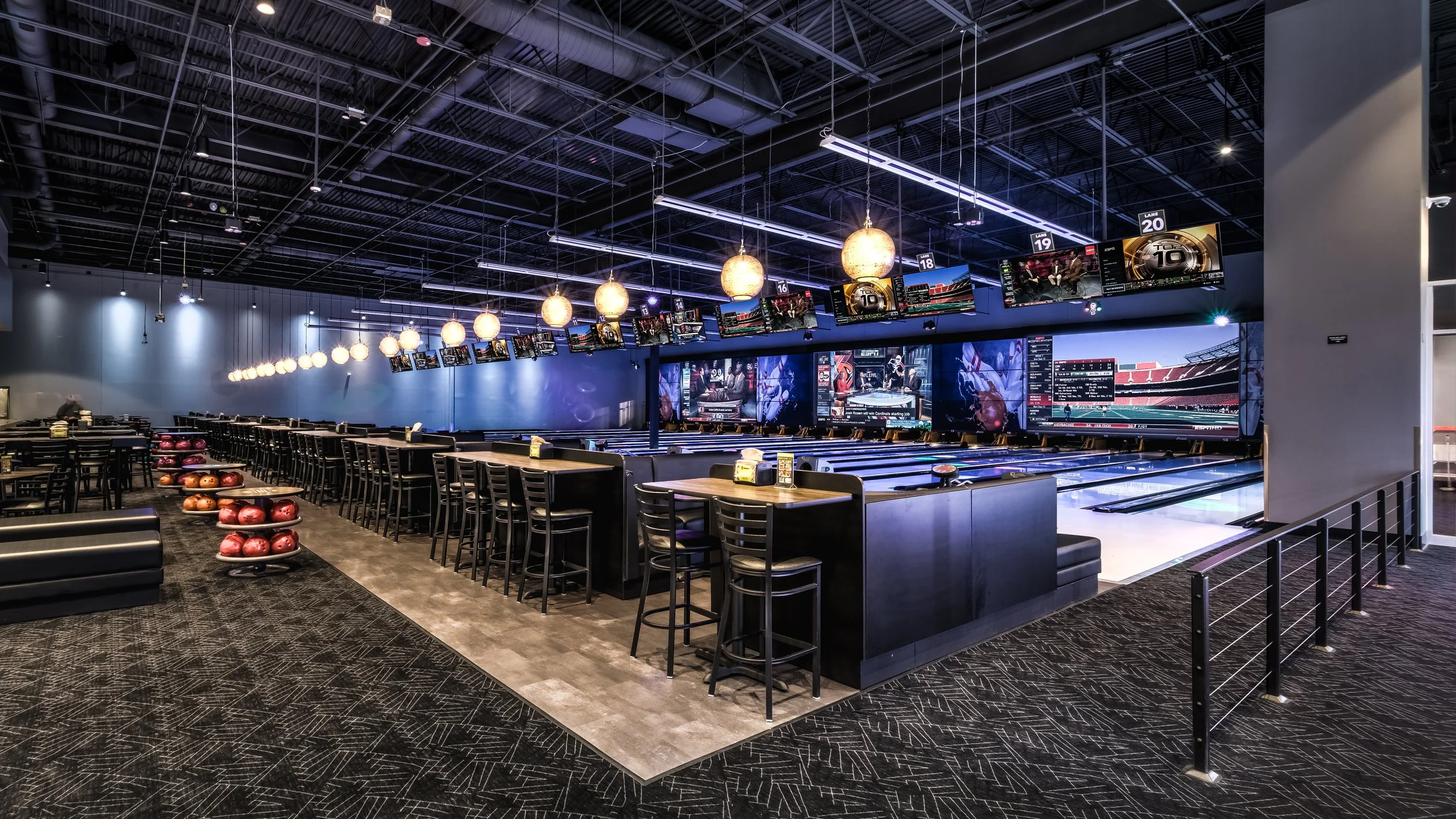 A modern bowling alley with multiple lanes, overhead monitors displaying game scores, and seating areas with tables and chairs.