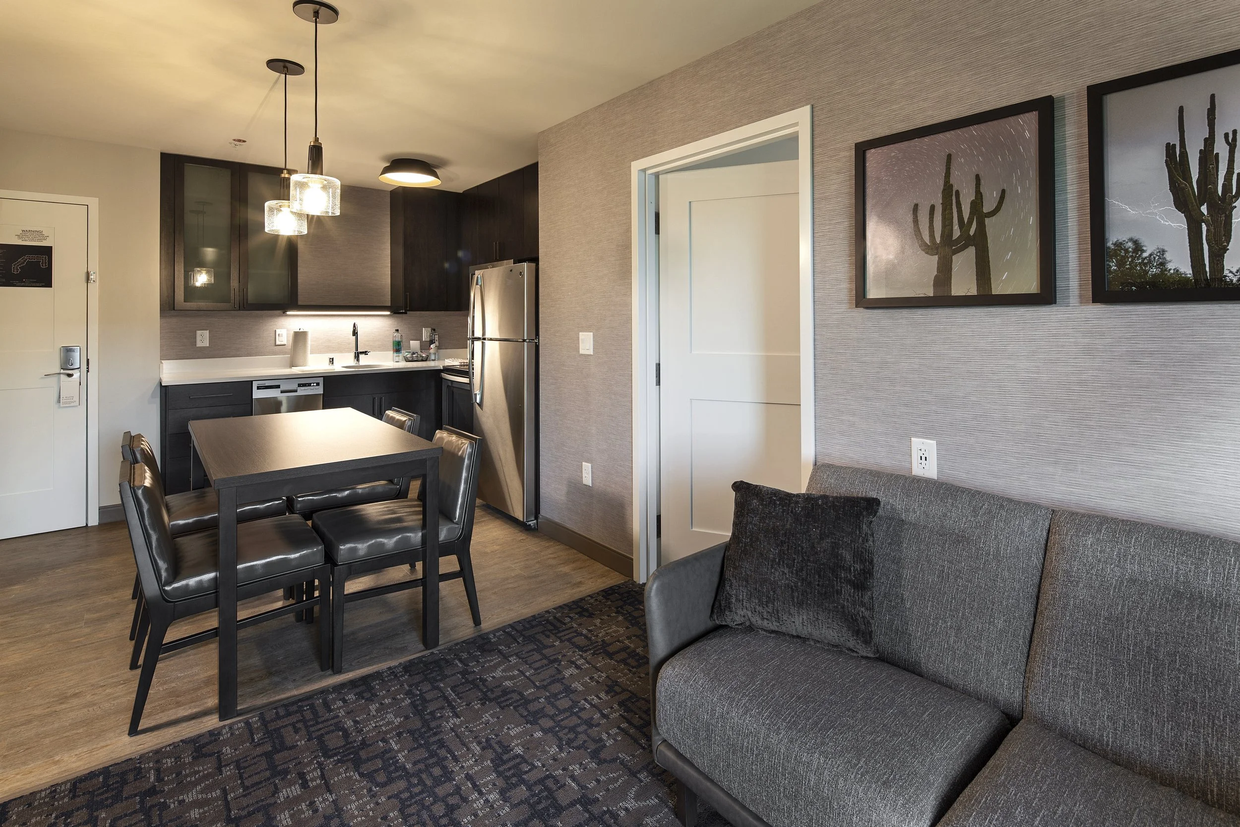 Modern kitchen and living area with dark cabinetry, stainless steel refrigerator, dining table with black chairs, gray couch, and framed cactus artwork on textured wall.