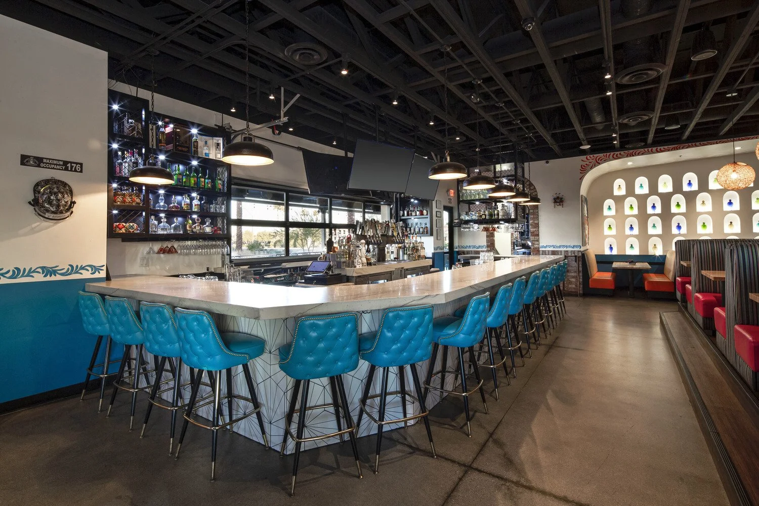 Interior of a modern bar with a white marble counter, blue barstools, and a lit bottle display behind the bar, with additional seating booths along the wall.