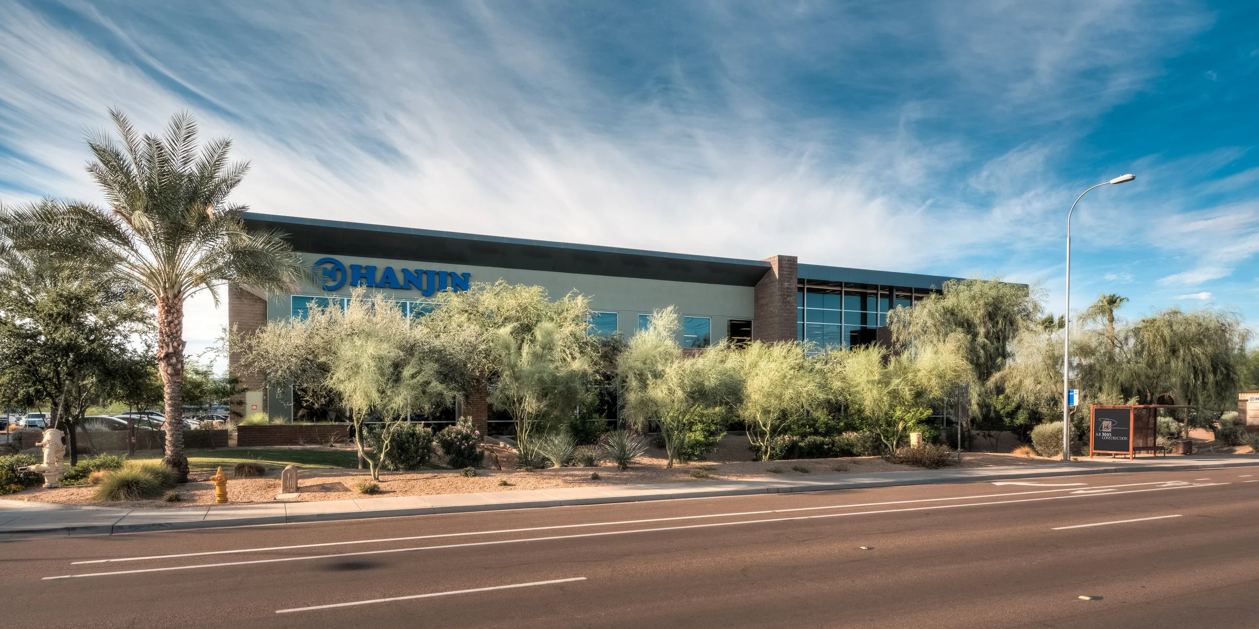 A modern commercial building with blue signage reading 'HANJIN', surrounded by desert landscaping and palm trees, under a partly cloudy sky along a multi-lane street.