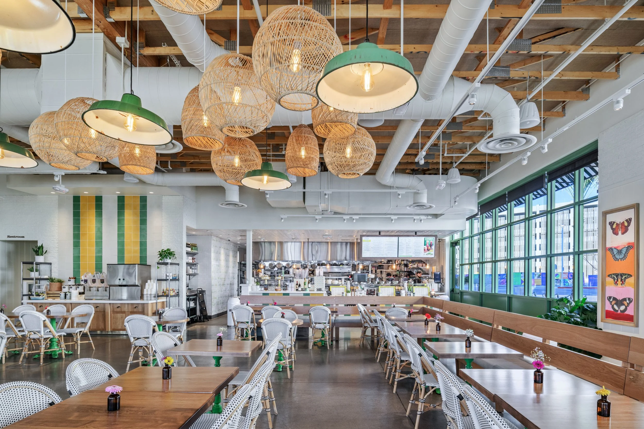 Interior of a modern cafe with hanging wicker and green lamps, wooden ceiling beams, large windows with black frames, tables with small vases of flowers, and a view of the kitchen and menu screens in the background.