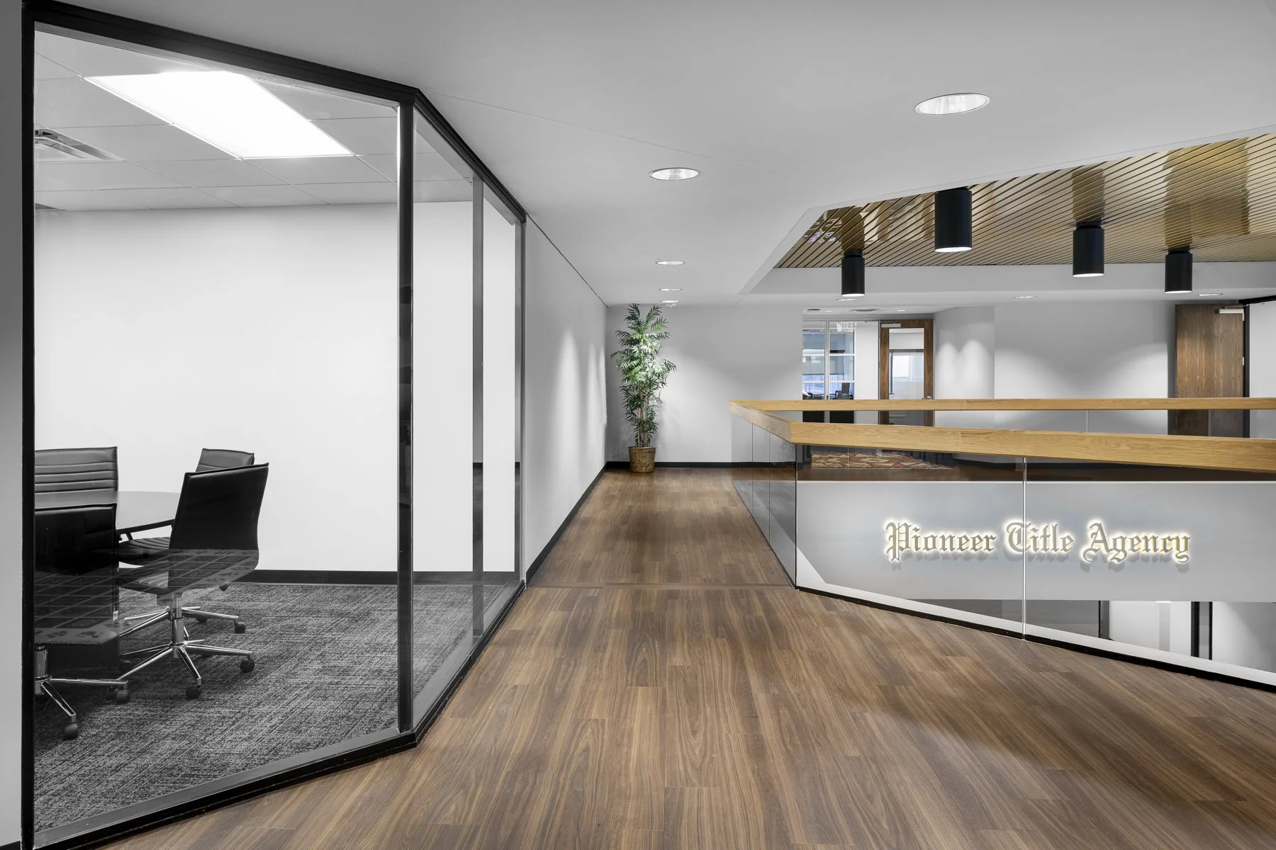 Office reception area with a glass-walled conference room on the left, wooden flooring, a reception desk that reads "Pioneer Title Agency," a potted plant, and modern lighting fixtures.