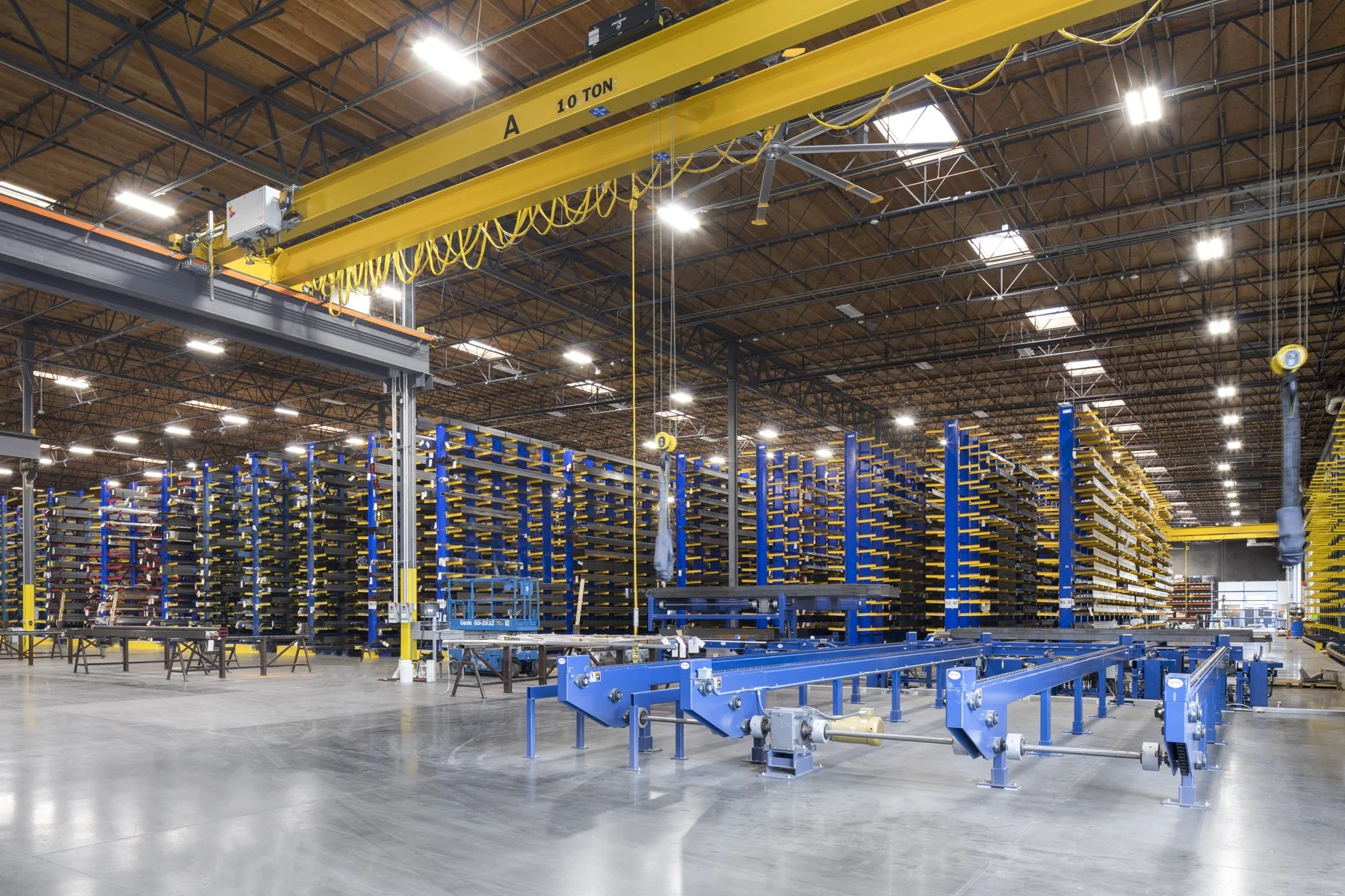 Inside a large warehouse with yellow overhead cranes, blue racks filled with yellow and black items, and a conveyor system on a smooth concrete floor.