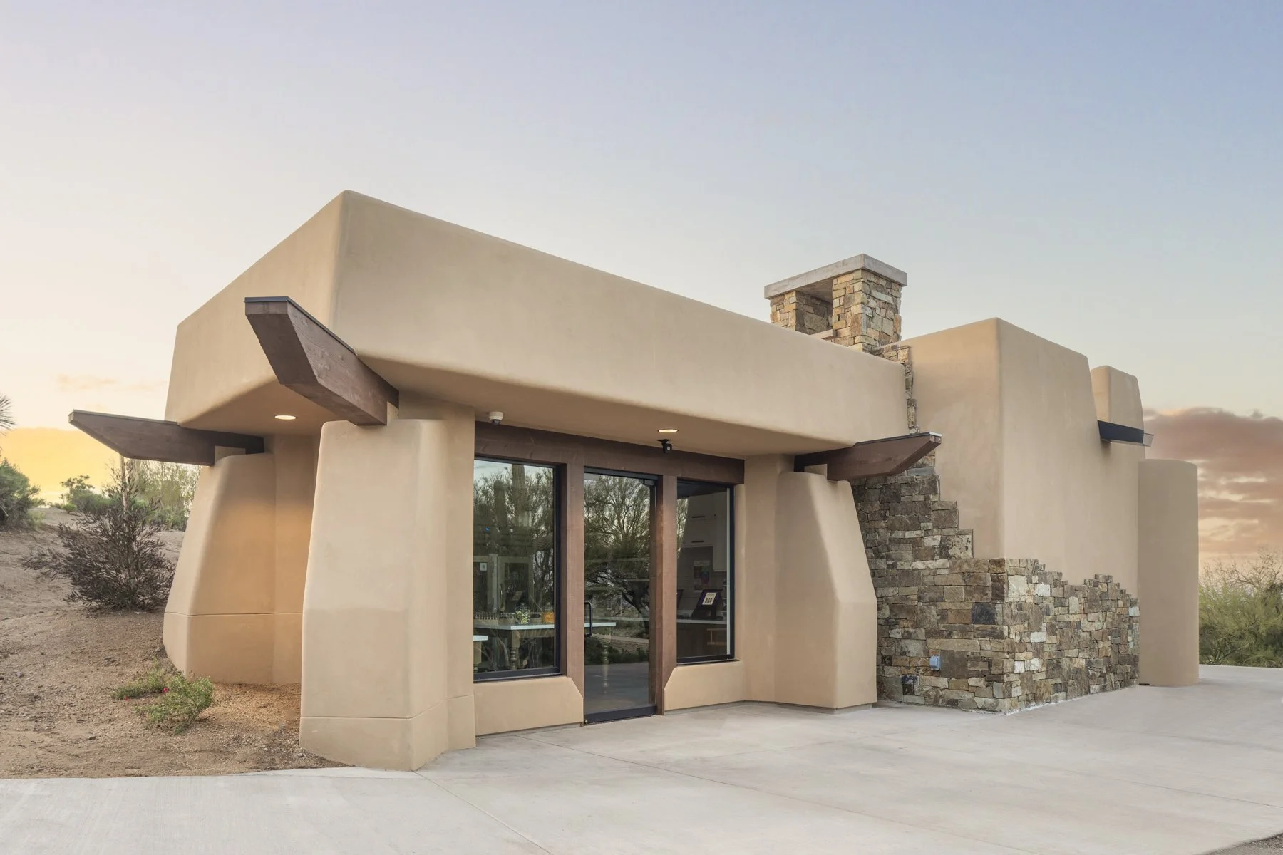 Modern southwestern style building with stucco walls, wooden beams, and a stone chimney, situated in a desert landscape at sunset.