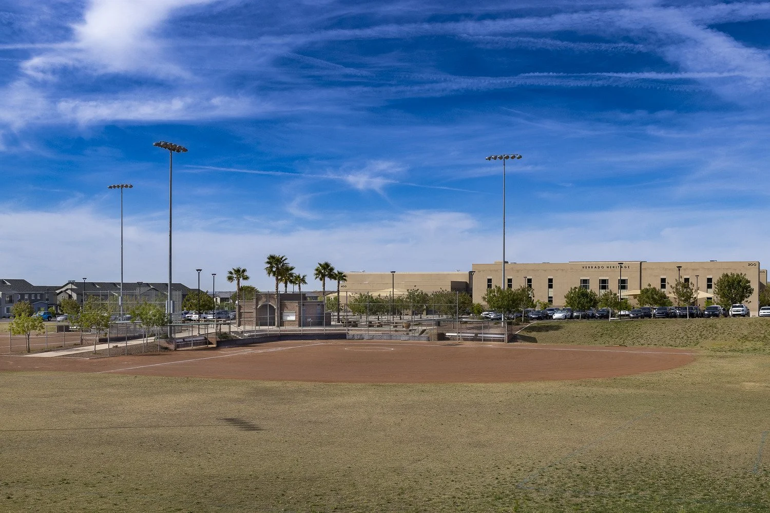 An empty baseball field with a dirt infield and green grass outfield, surrounded by a chain-link fence, with parking lot and trees in the background, under a blue sky with wispy clouds.