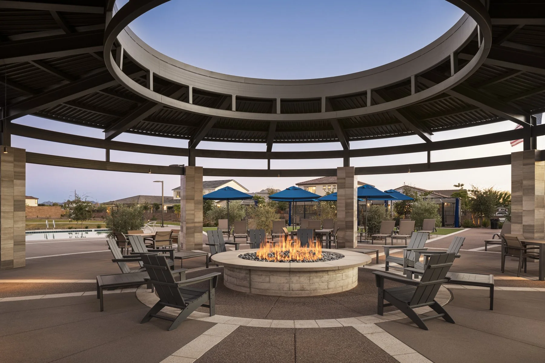 Outdoor communal area with a central fire pit surrounded by chairs, shaded by a circular open-roof structure with blue umbrellas and a swimming pool in the background.