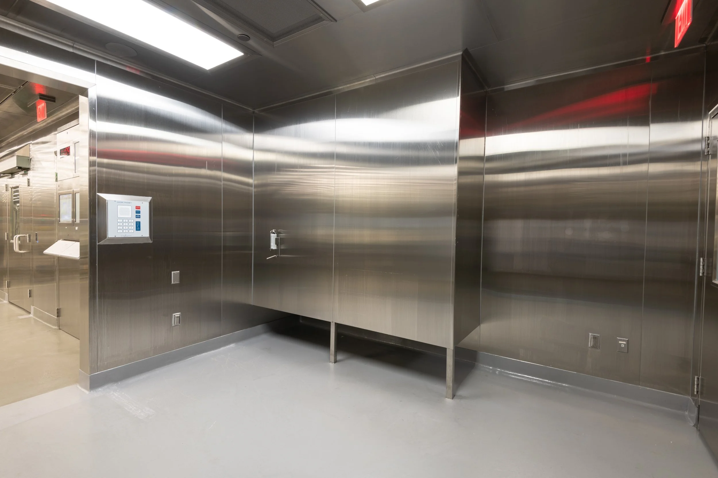 An empty room with stainless steel walls, a control panel, and a sink, likely a laboratory or clinical space.